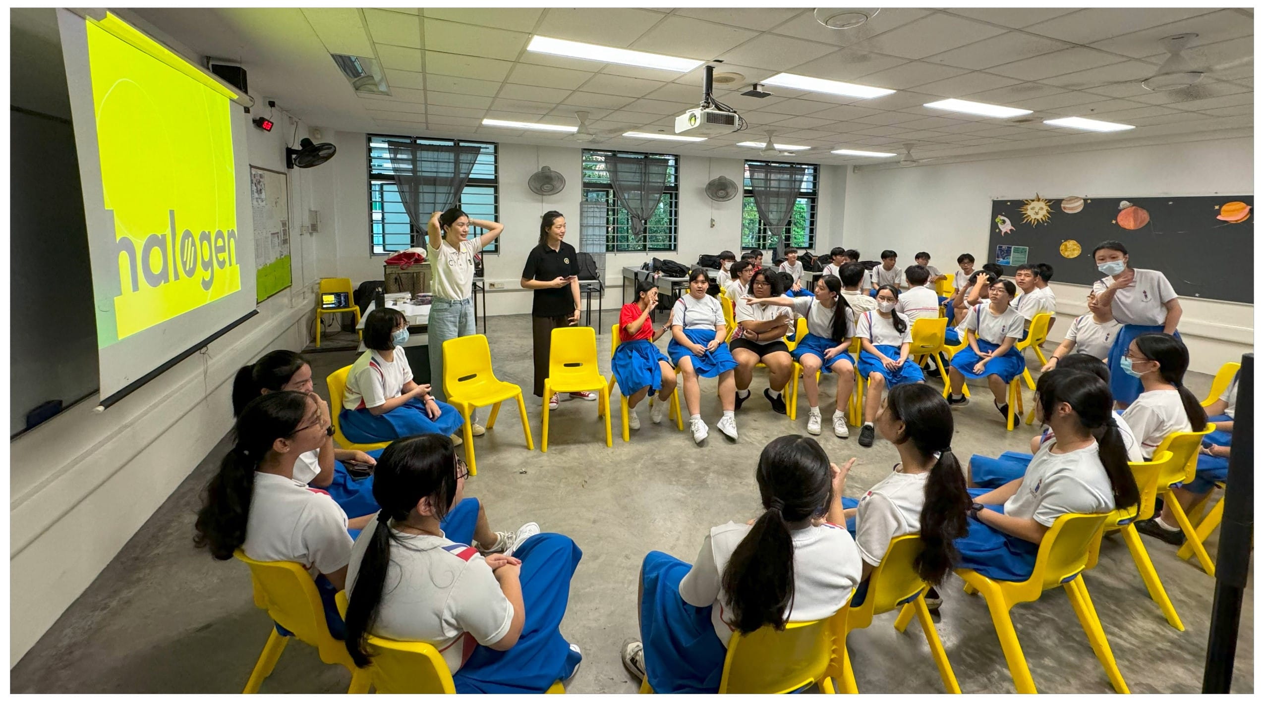 Students in uniforms sit in a circle on yellow chairs in a classroom with a bright yellow "halogen" slide projected.