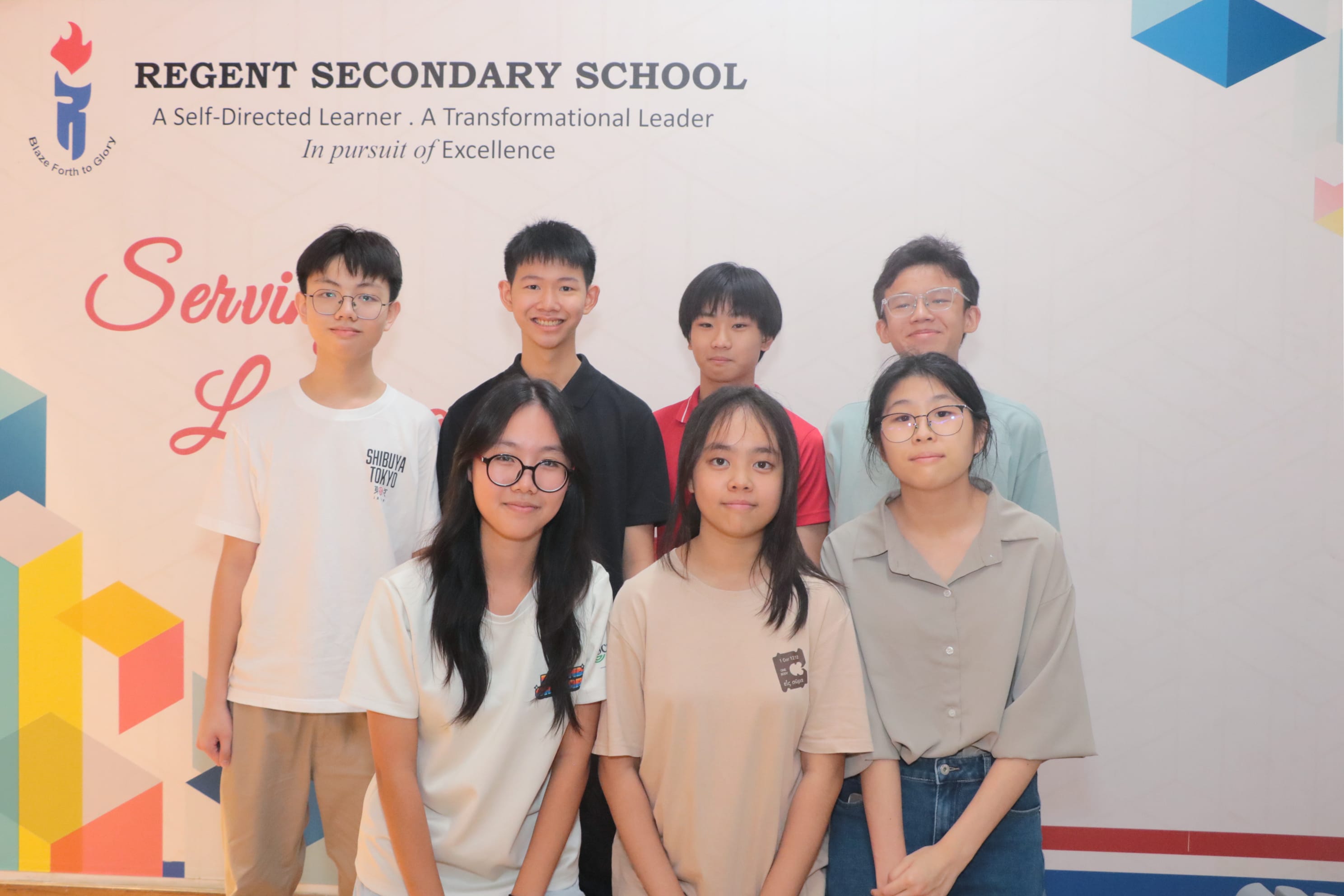 Seven students pose in two rows before a Regent Secondary School banner with colorful geometric decorations.