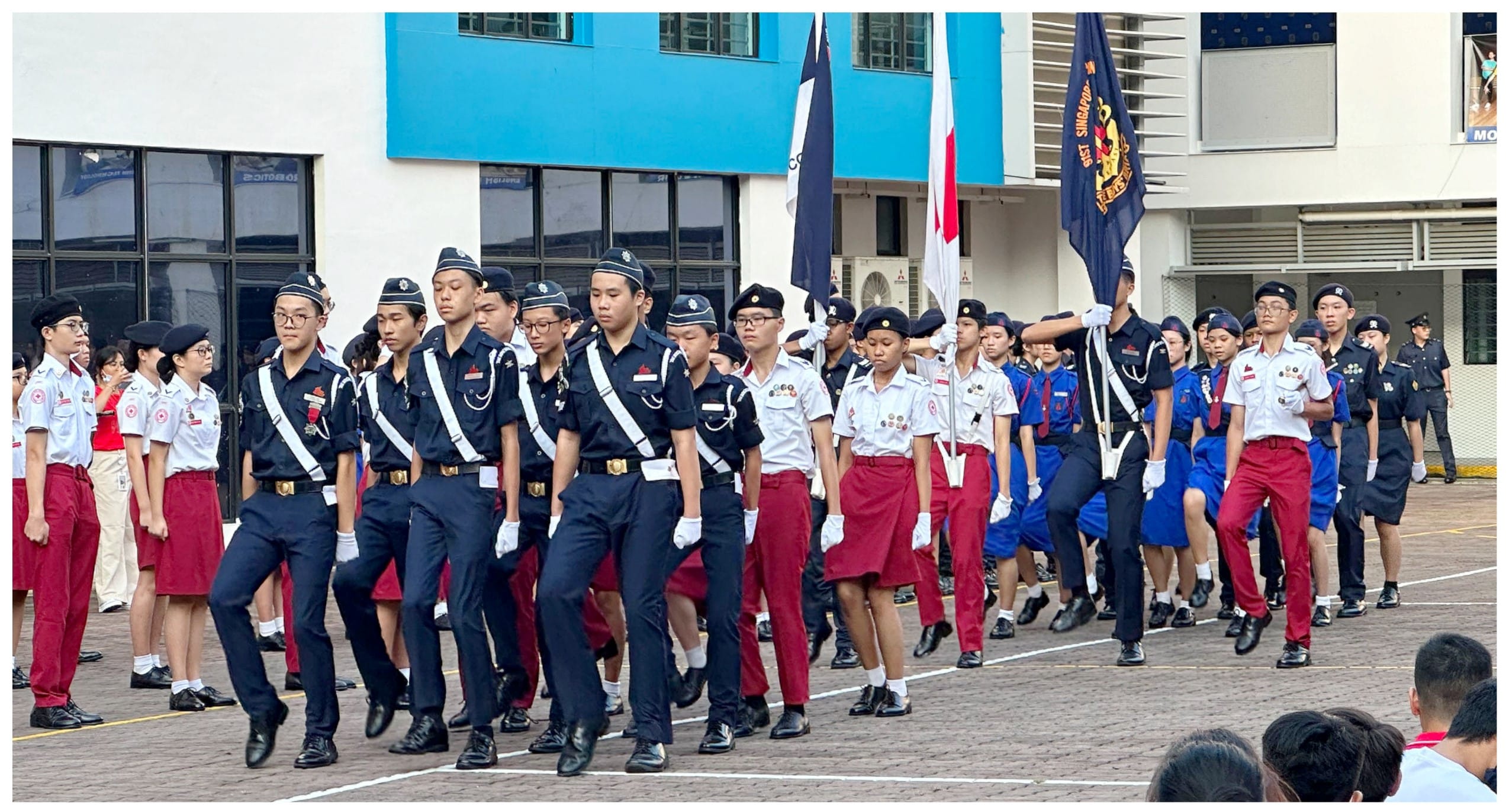 Students in various uniforms march in a formal parade on a school courtyard, carrying flags.