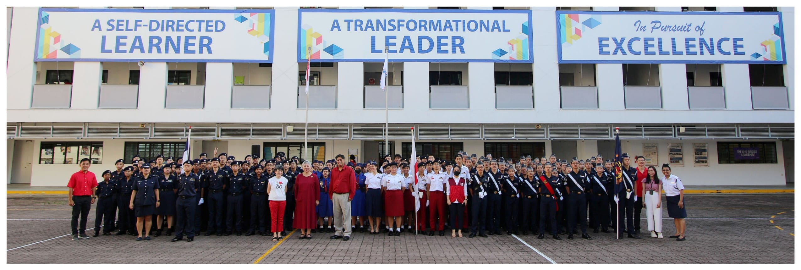Large group of uniformed students and staff posing outdoors in front of a school building with motivational banners.