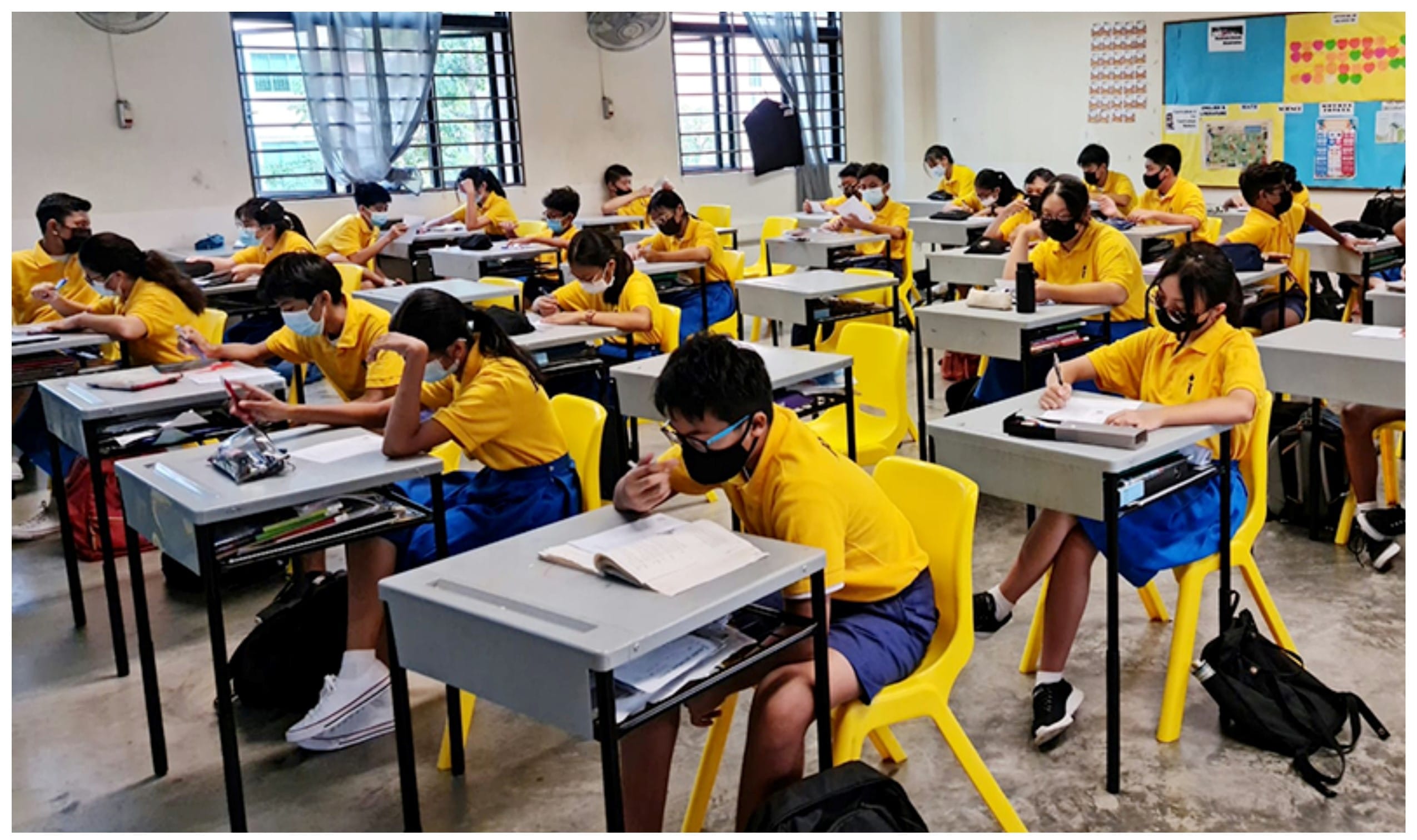 Students in yellow uniforms and face masks sit at desks writing in a full classroom with yellow chairs.