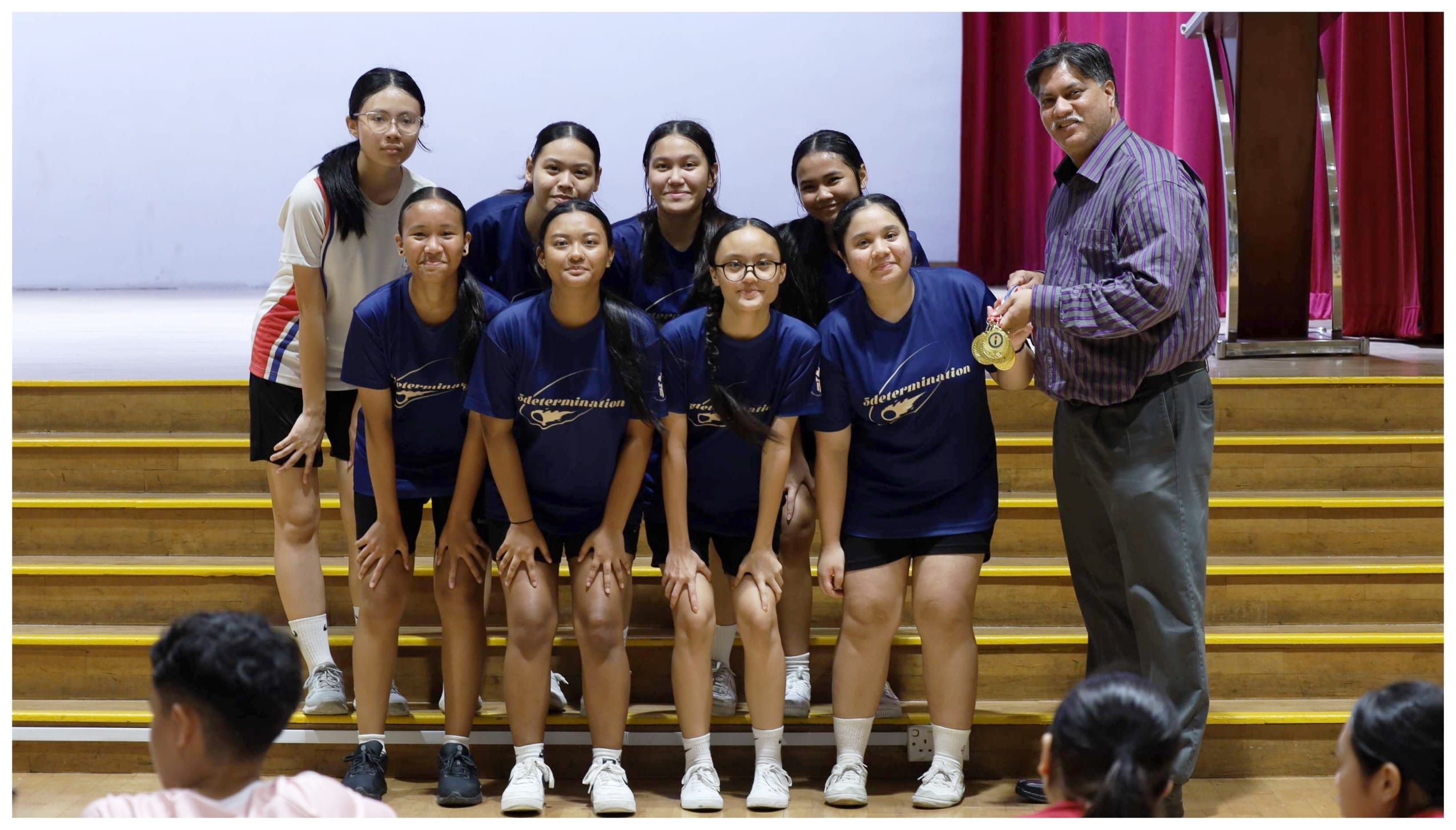 A man holds a gold medal beside a group of girls in matching blue sports jerseys posing on wooden bleachers.