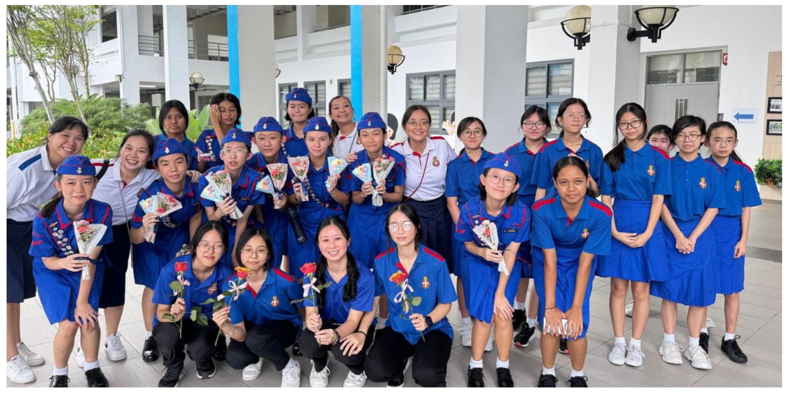 A large group of girls in blue uniforms holding flowers and bouquets, posing together outdoors near a building.