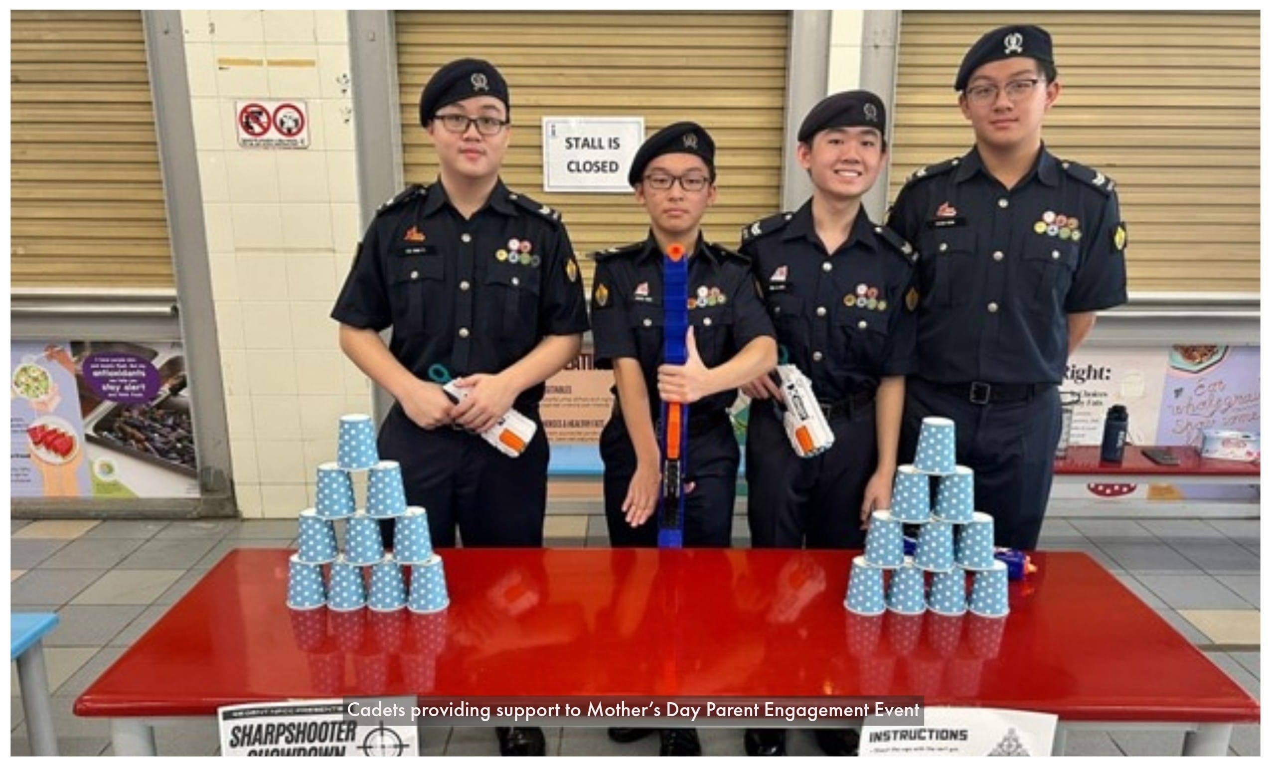 Four cadets in black uniforms hold toy guns beside two pyramid stacks of blue paper cups on a red table.