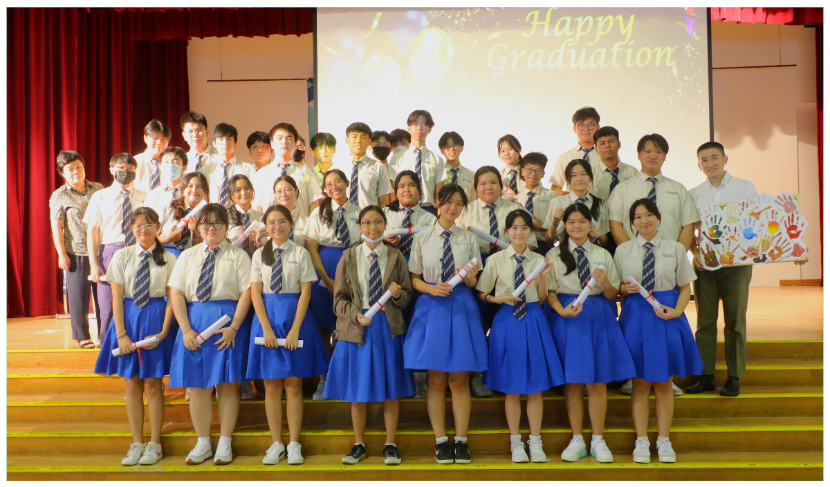 Students in uniforms pose on a stage holding diplomas, with a screen displaying "Happy Graduation" behind them.