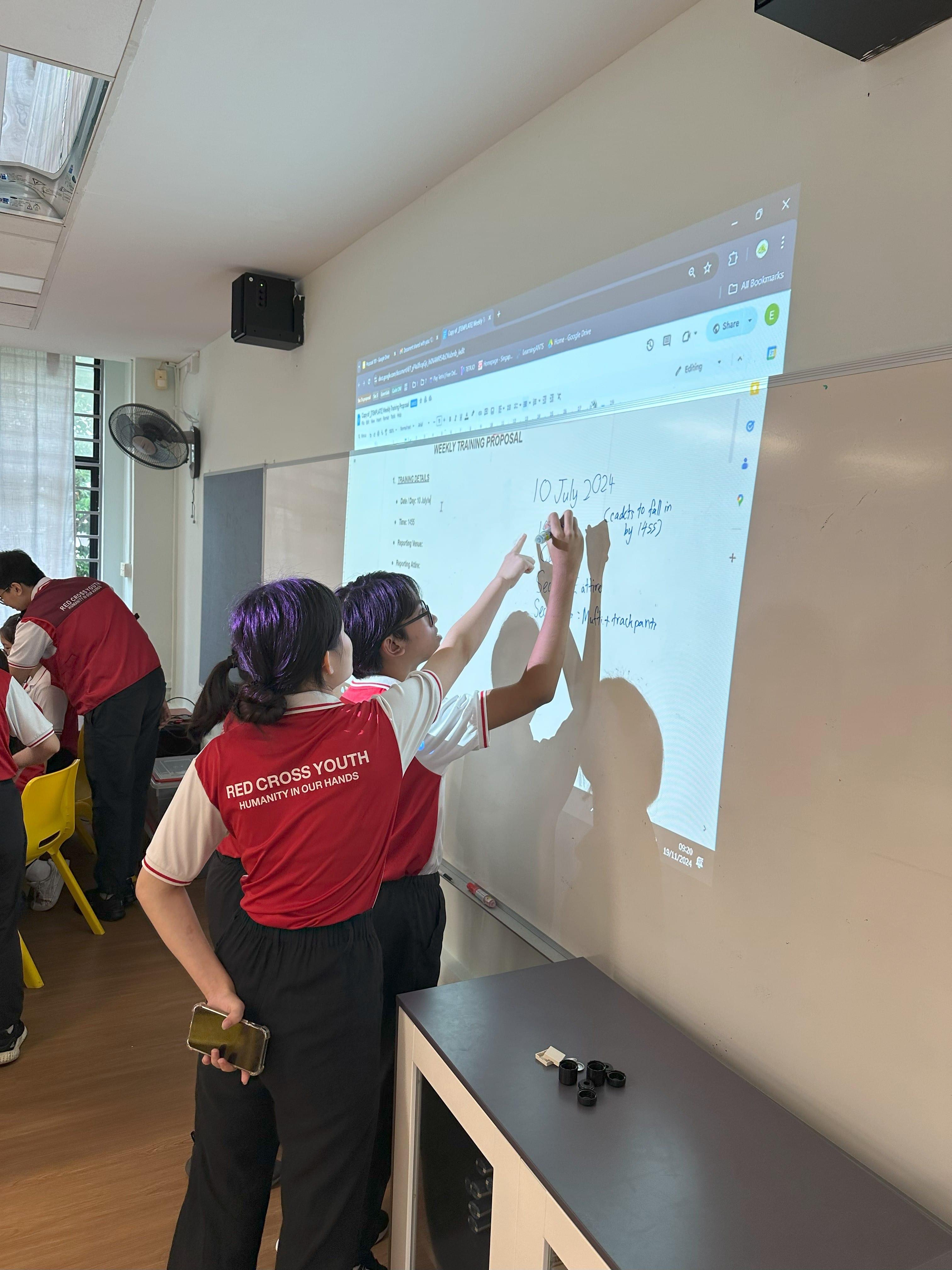 Two Red Cross Youth members in red uniforms write on a projected whiteboard showing a Weekly Training Proposal document.