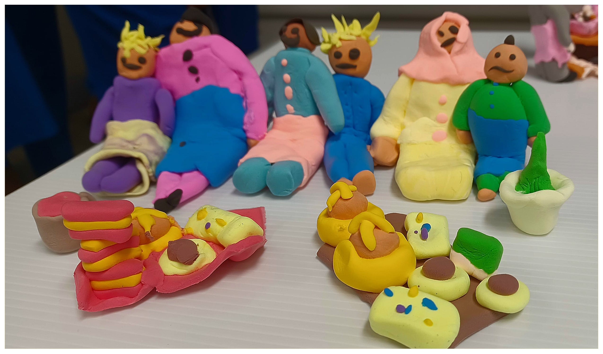 Colorful clay figurines of people in pink, blue, yellow, and green sit on a white table with small clay objects.