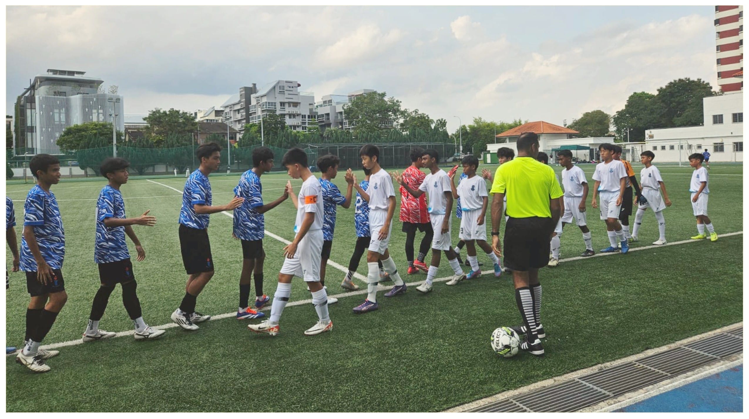Two youth soccer teams in blue and white uniforms shake hands on a green turf field, with a referee in yellow nearby.