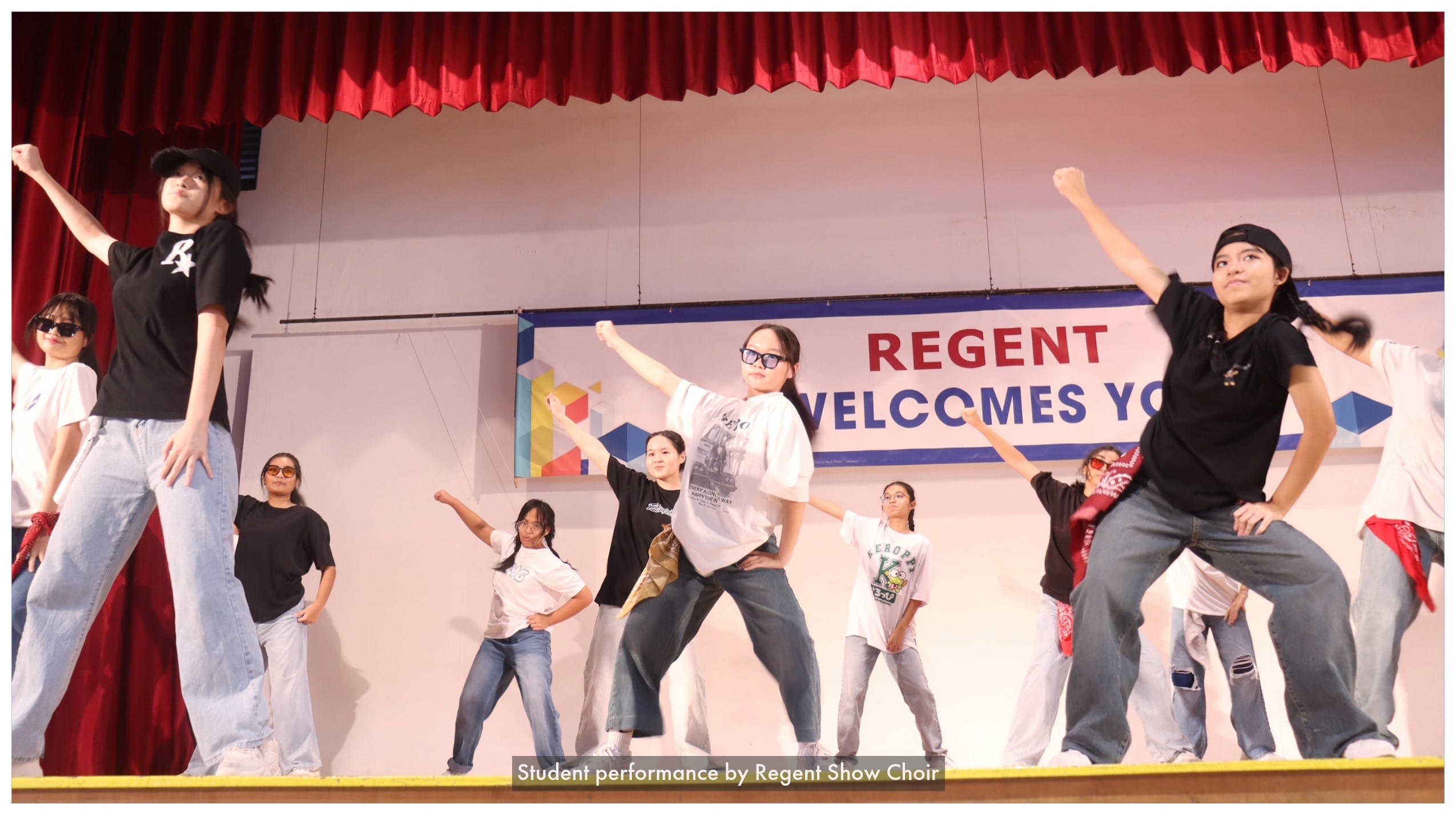 Students perform a hip-hop dance on stage beneath a red curtain and a Regent Welcomes You banner.