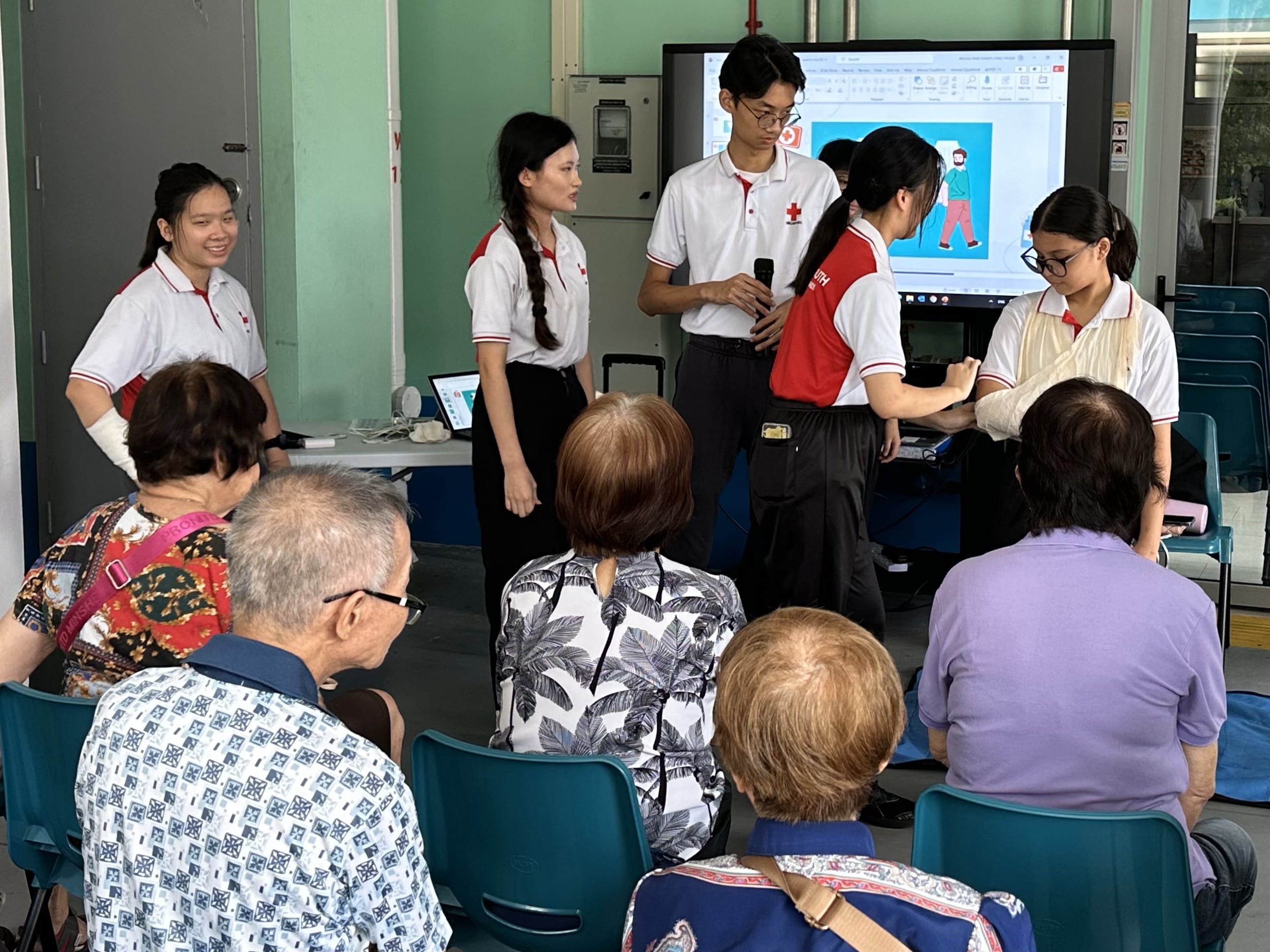 Red Cross youth volunteers present to a group of seated elderly people in front of a screen showing a health illustration.