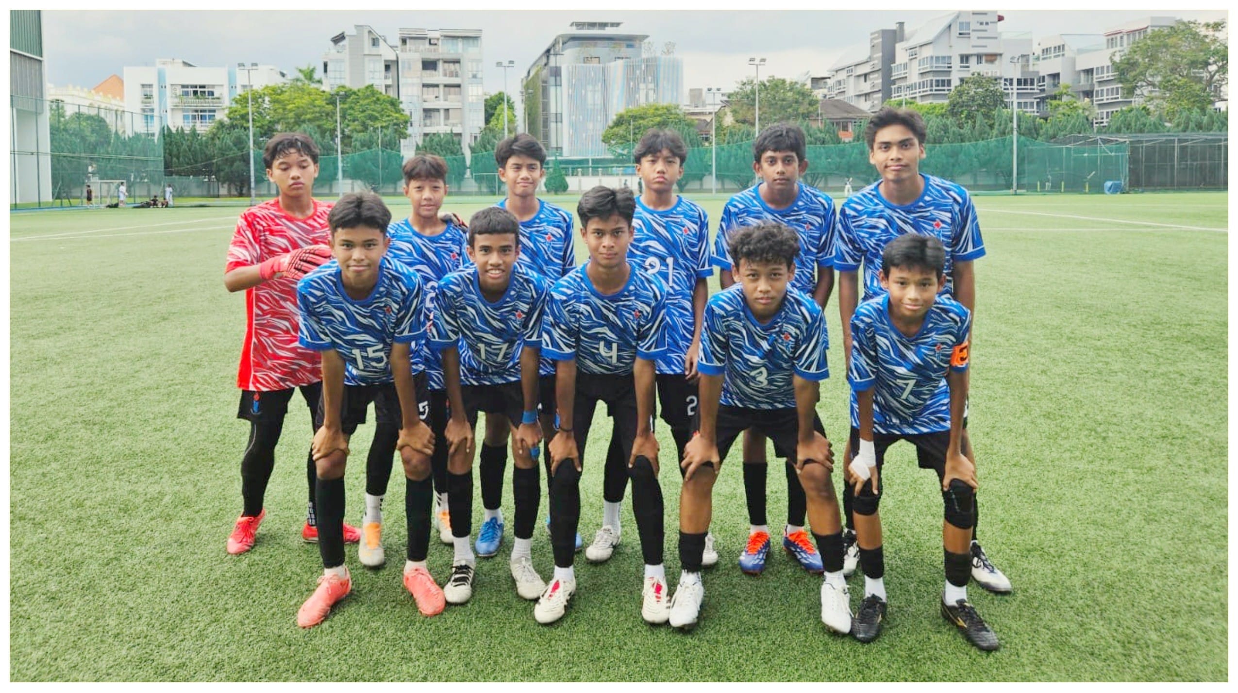 A youth football team of 11 boys in blue jerseys poses on a green field, with buildings and trees in the background.