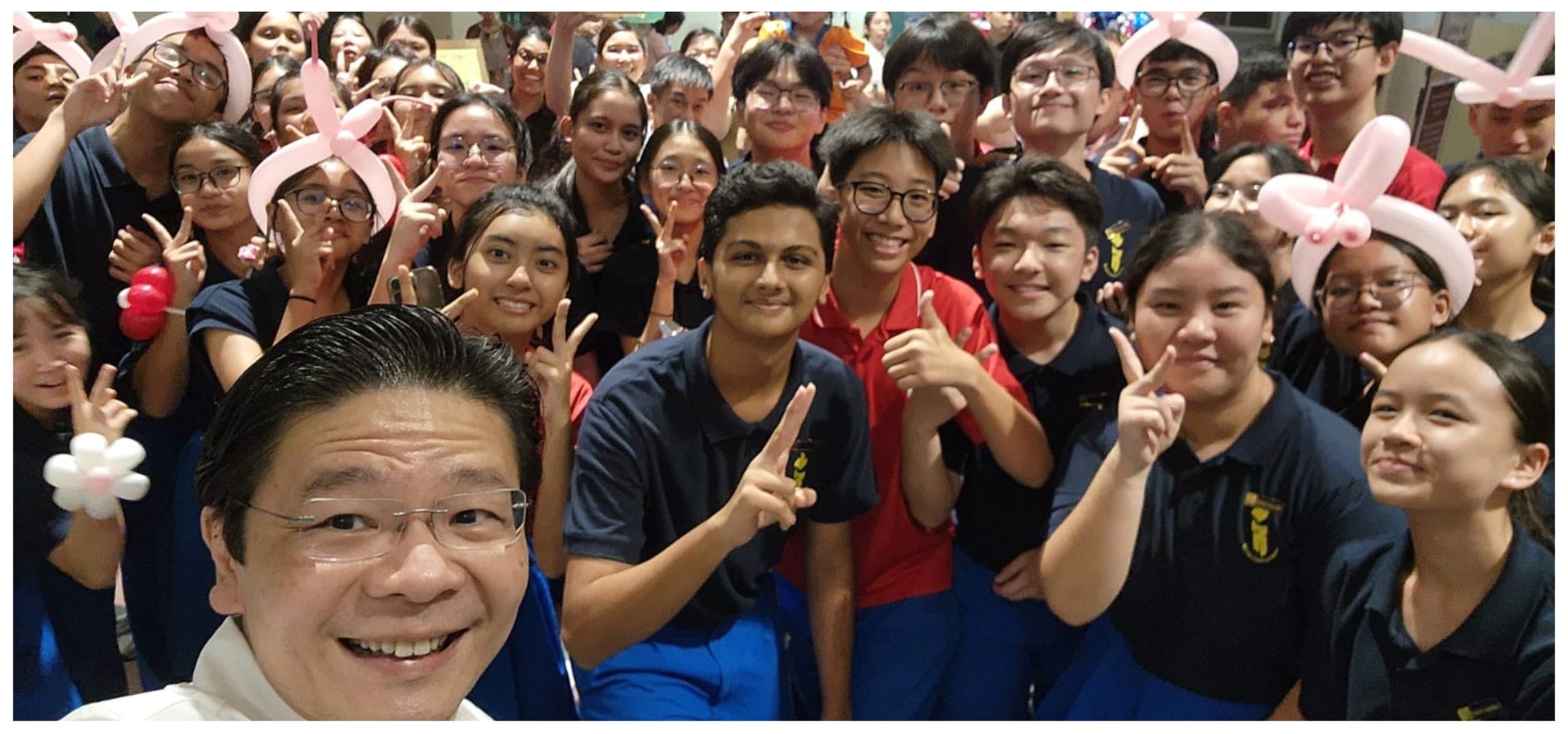 A large group of smiling students in navy uniforms pose with peace signs and pink balloon hats in a selfie.