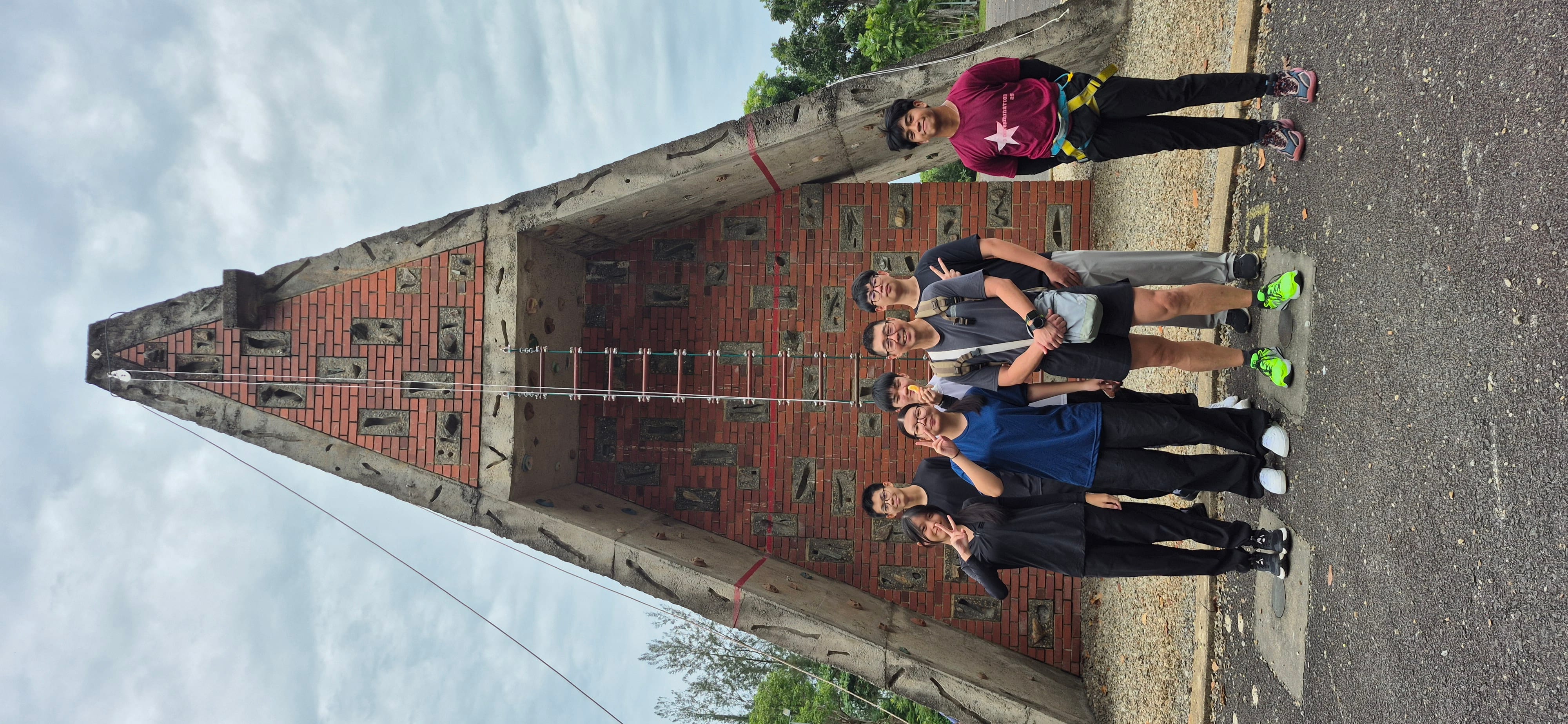 Six young people pose in front of a tall triangular brick climbing wall with a rope ladder under a cloudy sky.