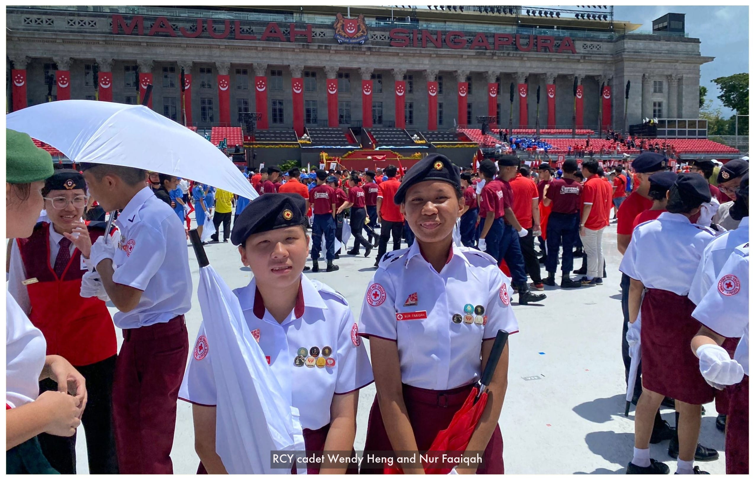 Two Red Cross Youth cadets in white uniforms and black berets stand at a Singapore National Day parade.