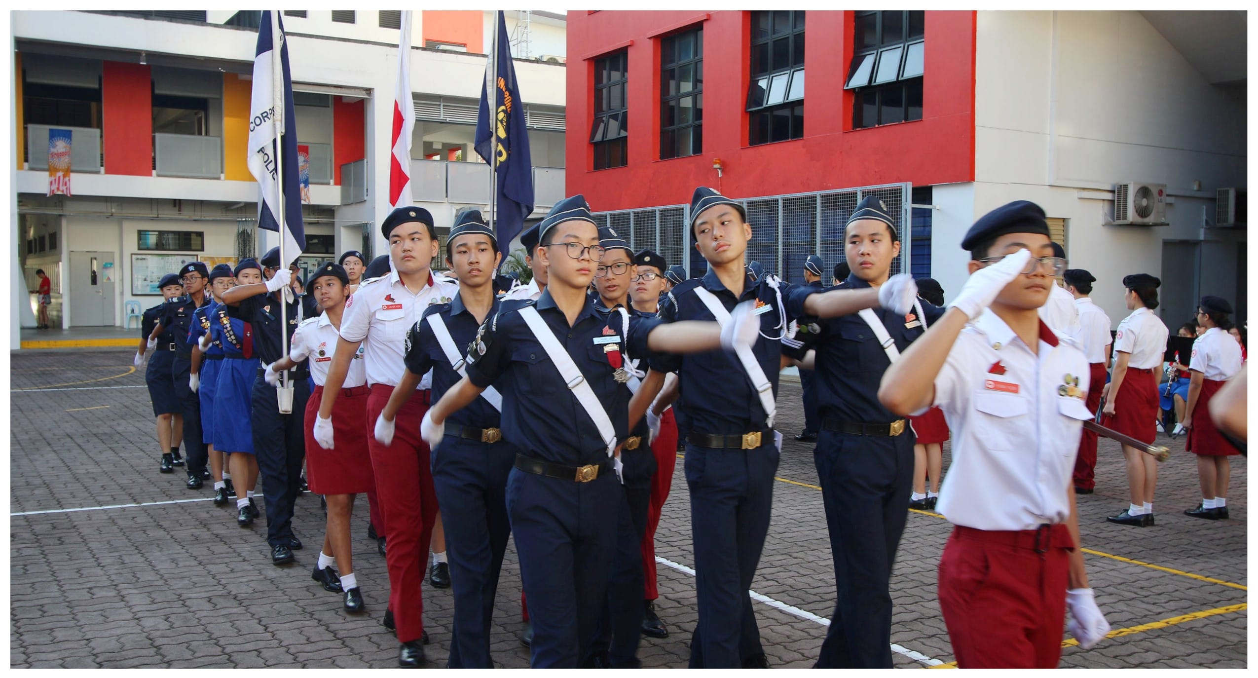 Students in navy uniforms and berets march in formation carrying flags during a school parade ceremony.