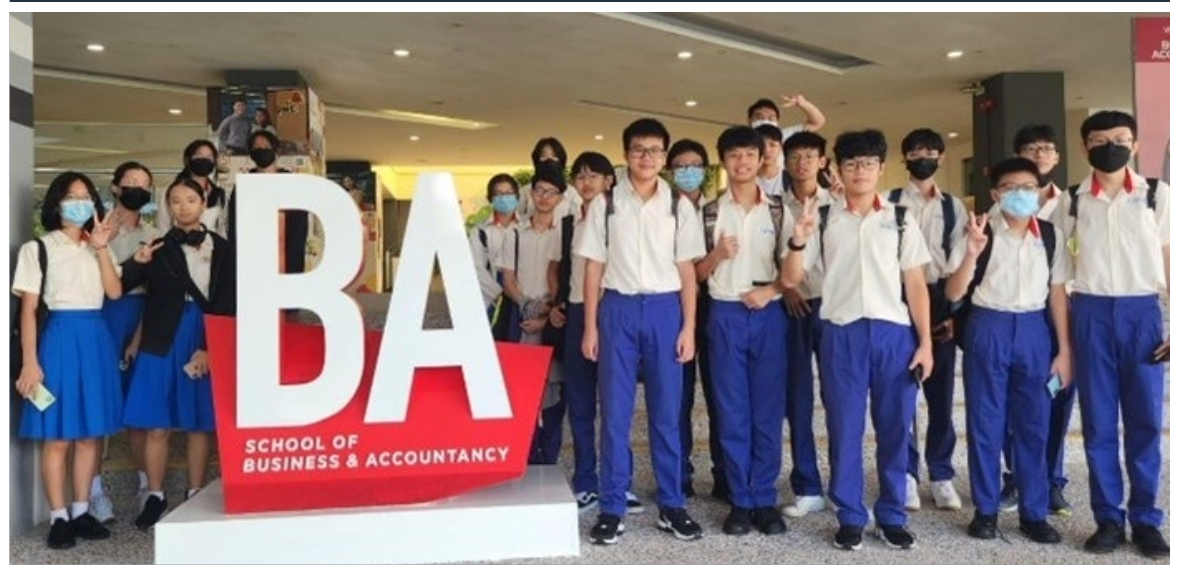 Students in school uniforms and face masks pose beside a large red BA School of Business and Accountancy sign.