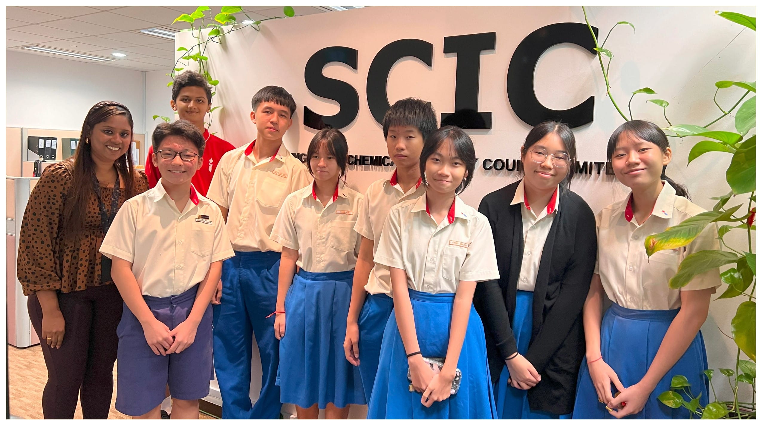 A group of students in white and blue uniforms pose with an adult woman in front of a wall with SCIC letters.