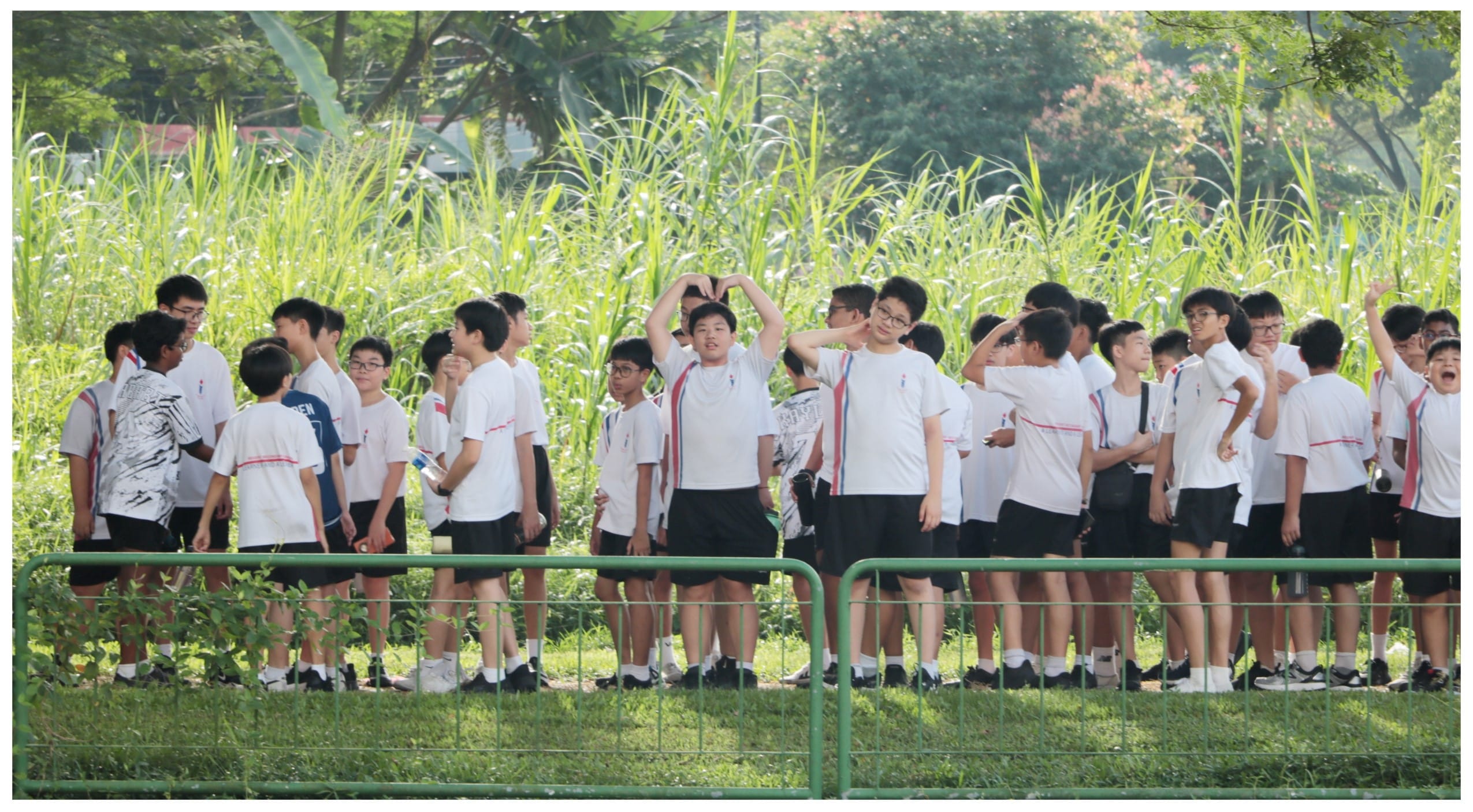A large group of boys in white shirts and black shorts stand outdoors near tall grass, one making a heart shape overhead.