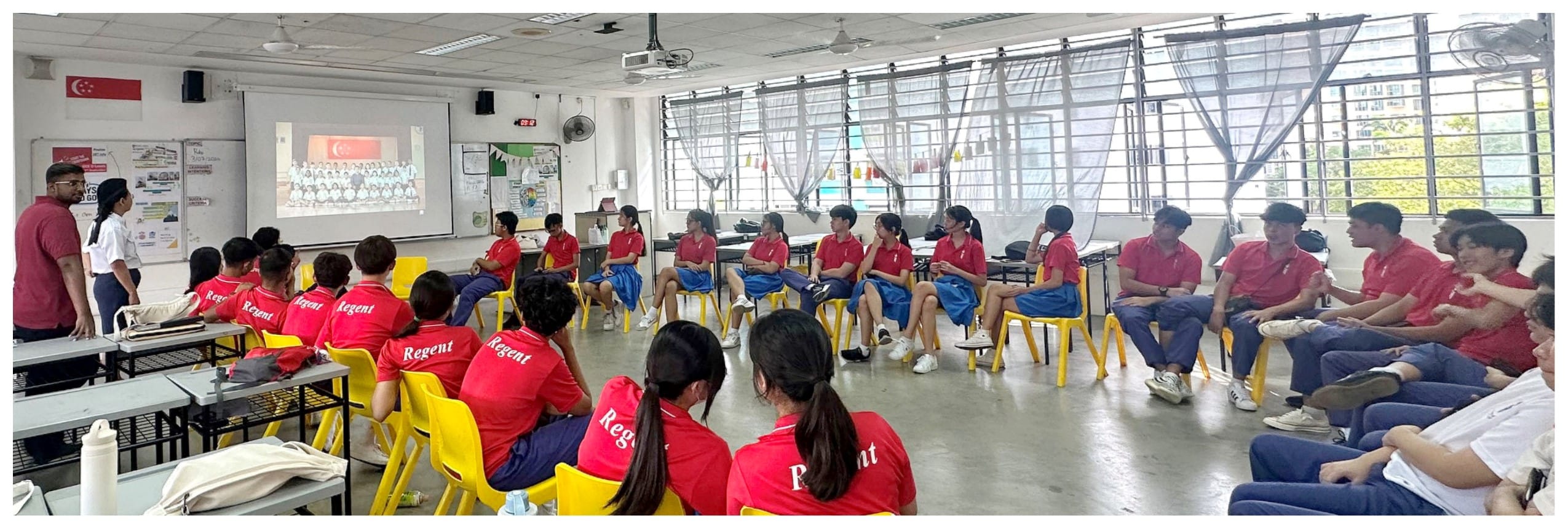 Students in red Regent uniforms sit in a circle in a classroom, watching a projected presentation on a whiteboard.