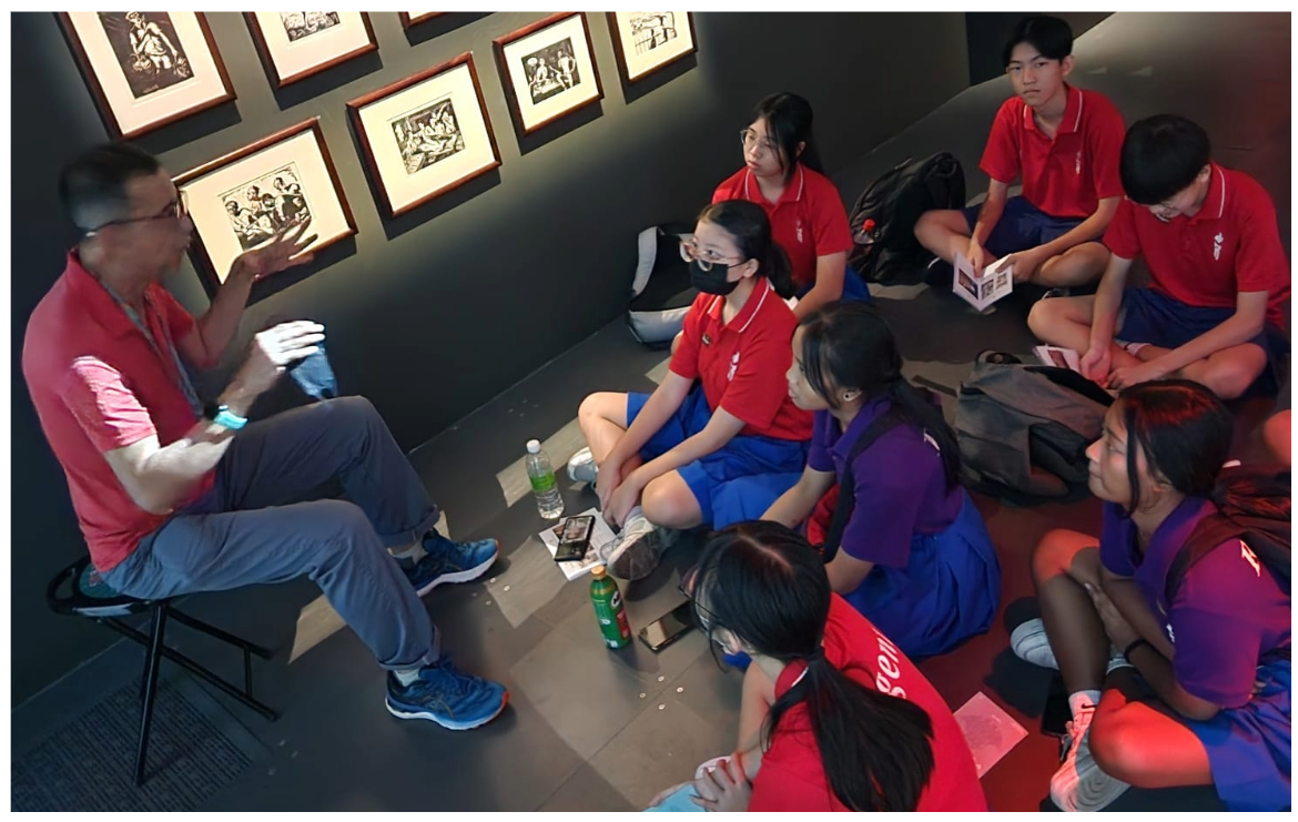 A man in a red shirt speaks to students in school uniforms seated on the floor of an art gallery.