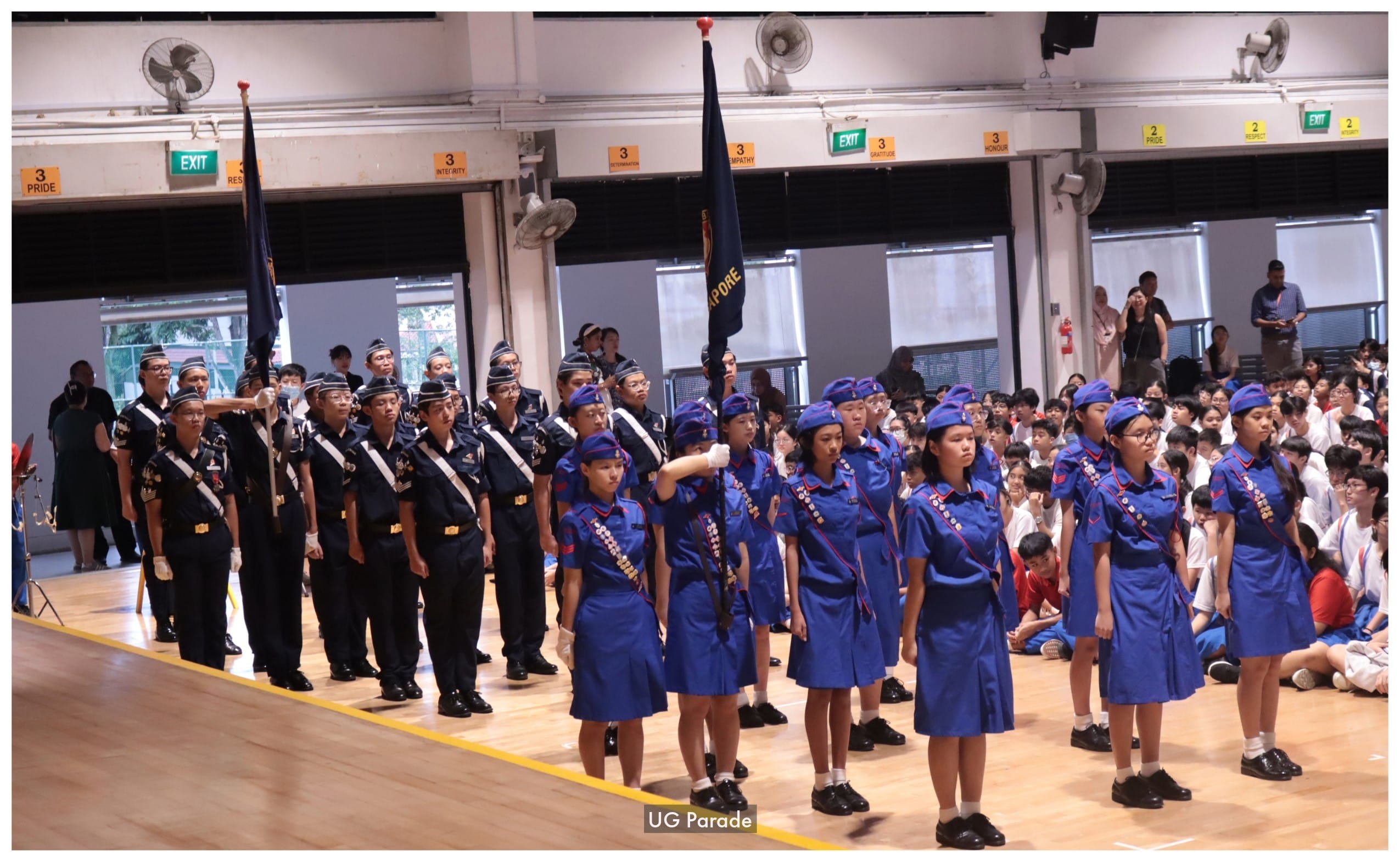 Students in navy and blue uniforms stand in formation holding flags during a school parade in an indoor hall.