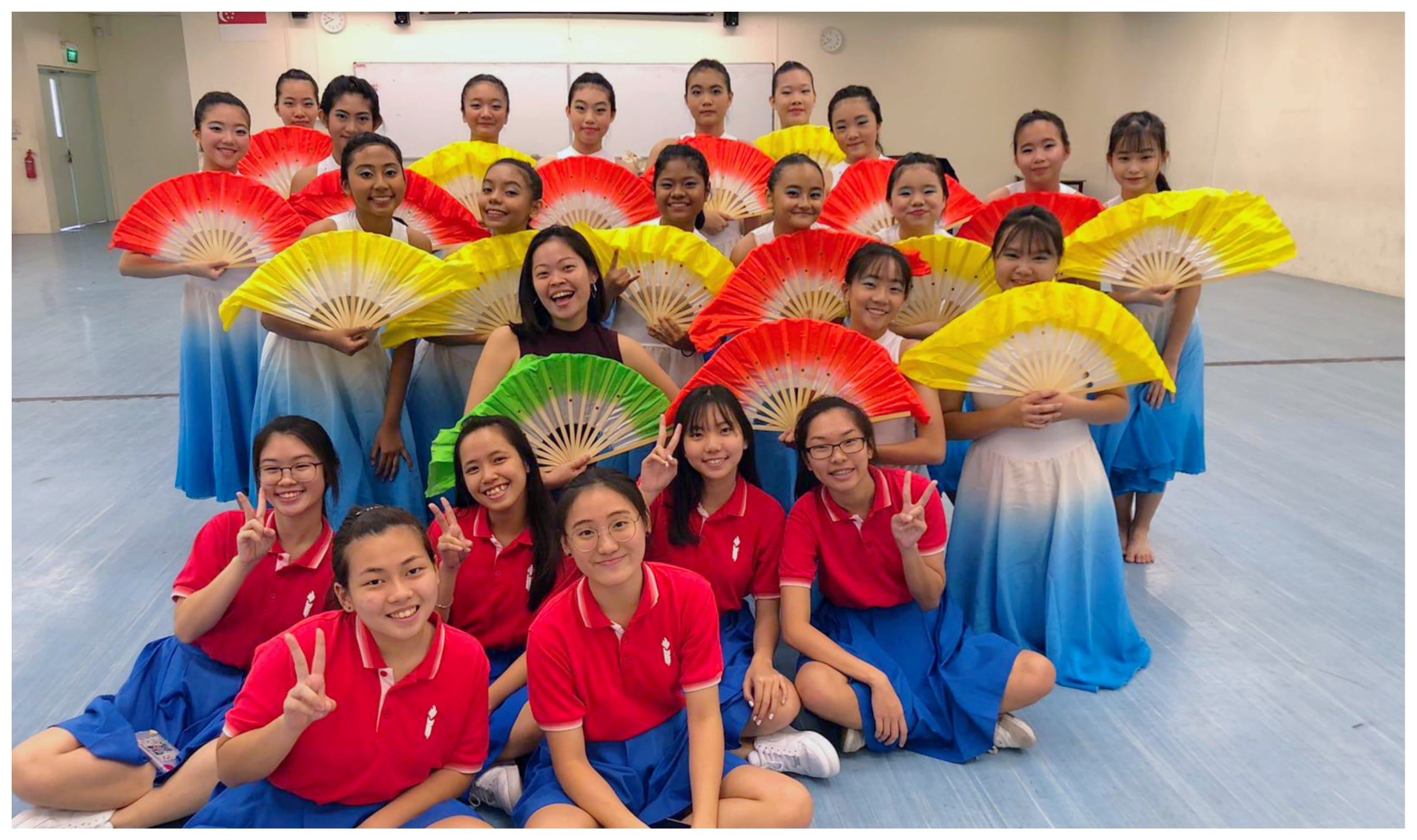 A large group of young women pose together, some holding colorful red and yellow fans, wearing white dresses or red uniforms.