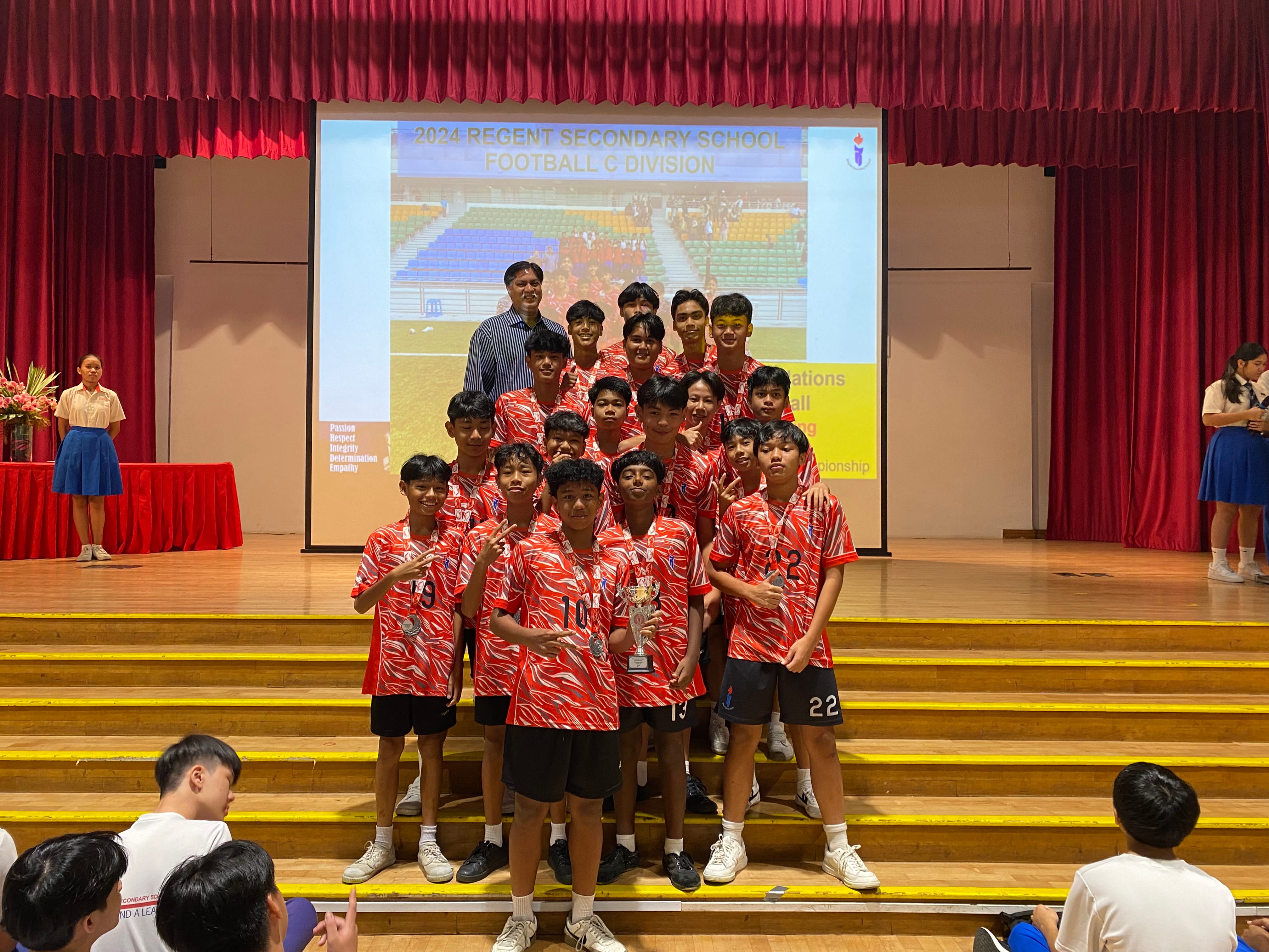 A youth football team in red jerseys poses on stage steps holding a trophy at a 2024 Regent Secondary School event.