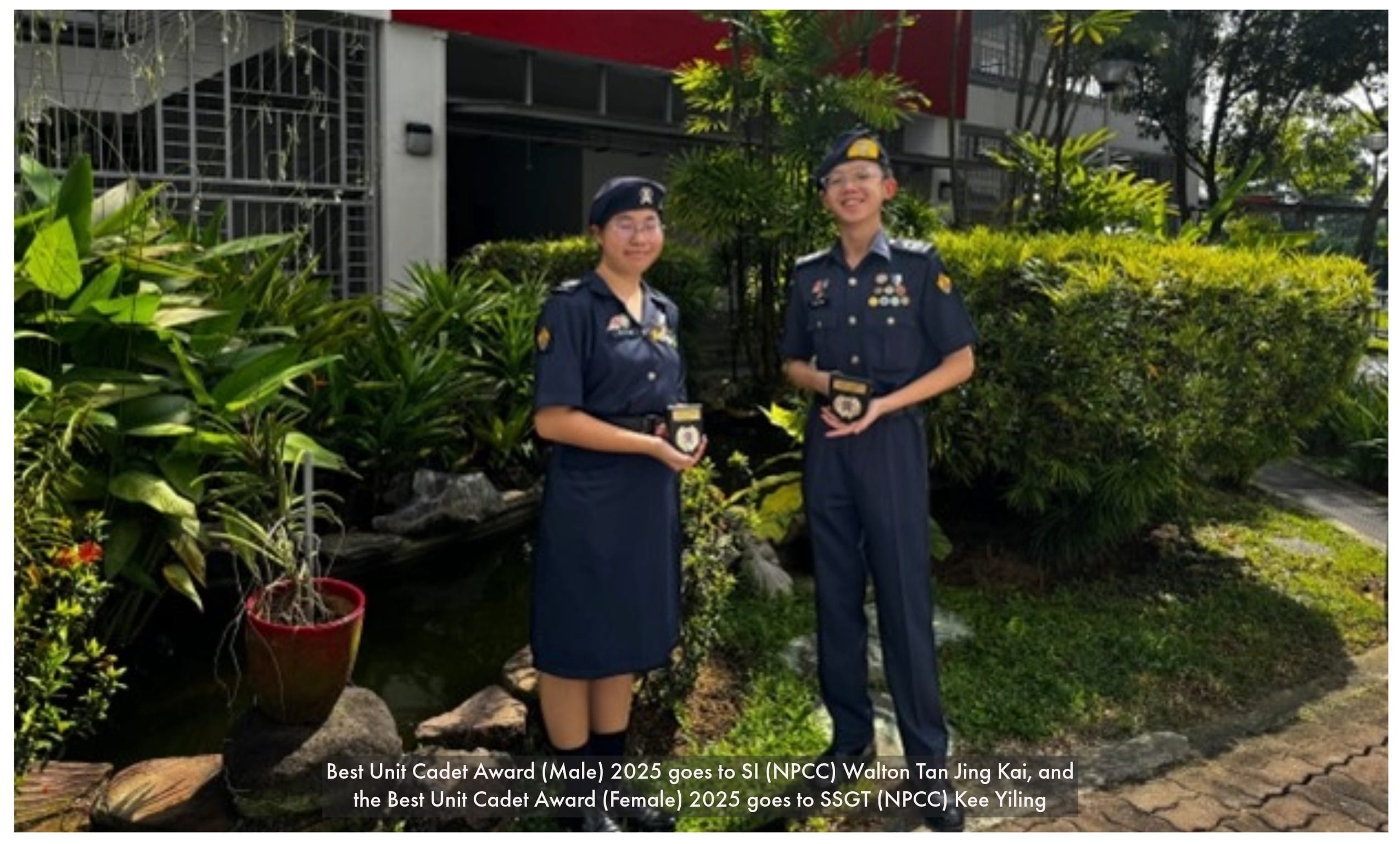 Two NPCC cadets in navy uniforms holding awards, standing outdoors among green plants and shrubs.