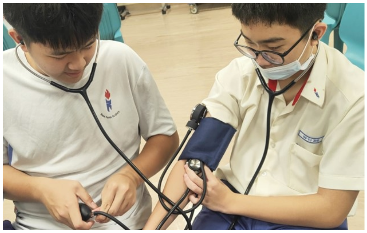 Two male students in uniforms practice taking blood pressure, one wearing a mask and glasses, both using stethoscopes.
