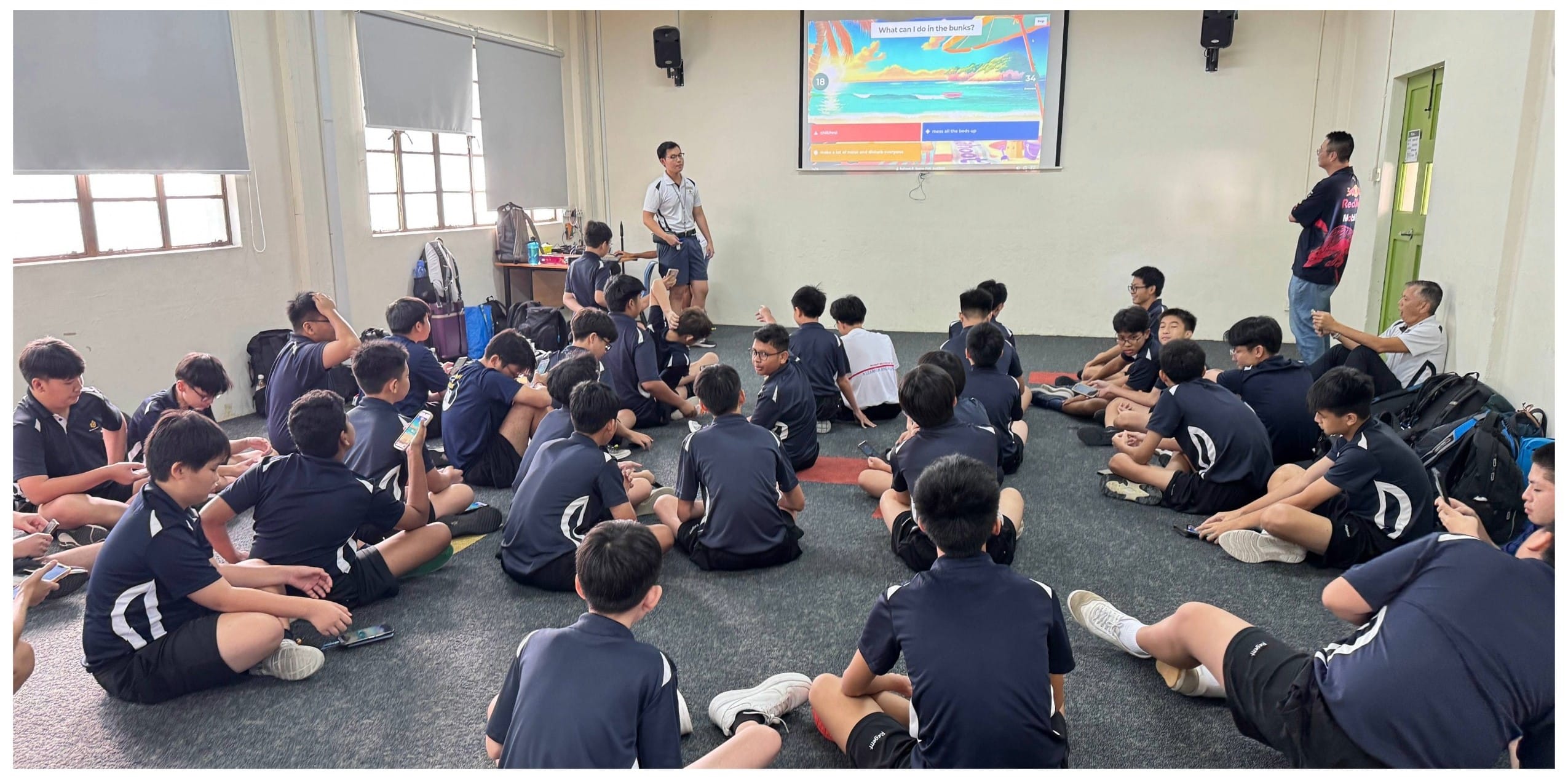 Students in navy uniforms sit on a floor facing a presenter and a projected quiz slide on a screen.