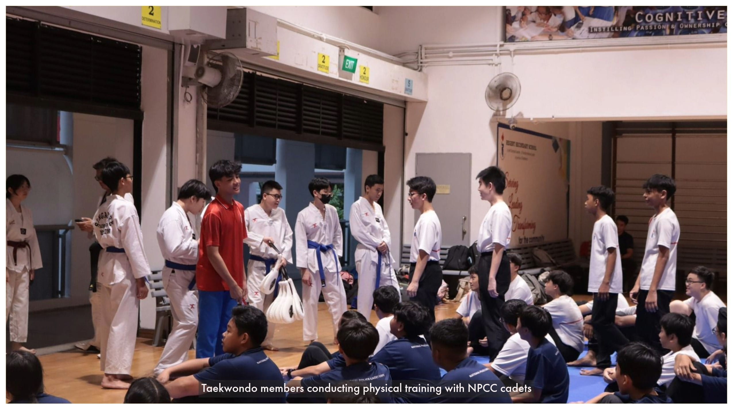 Students in white taekwondo uniforms with blue belts stand before seated cadets in navy uniforms inside a school hall.