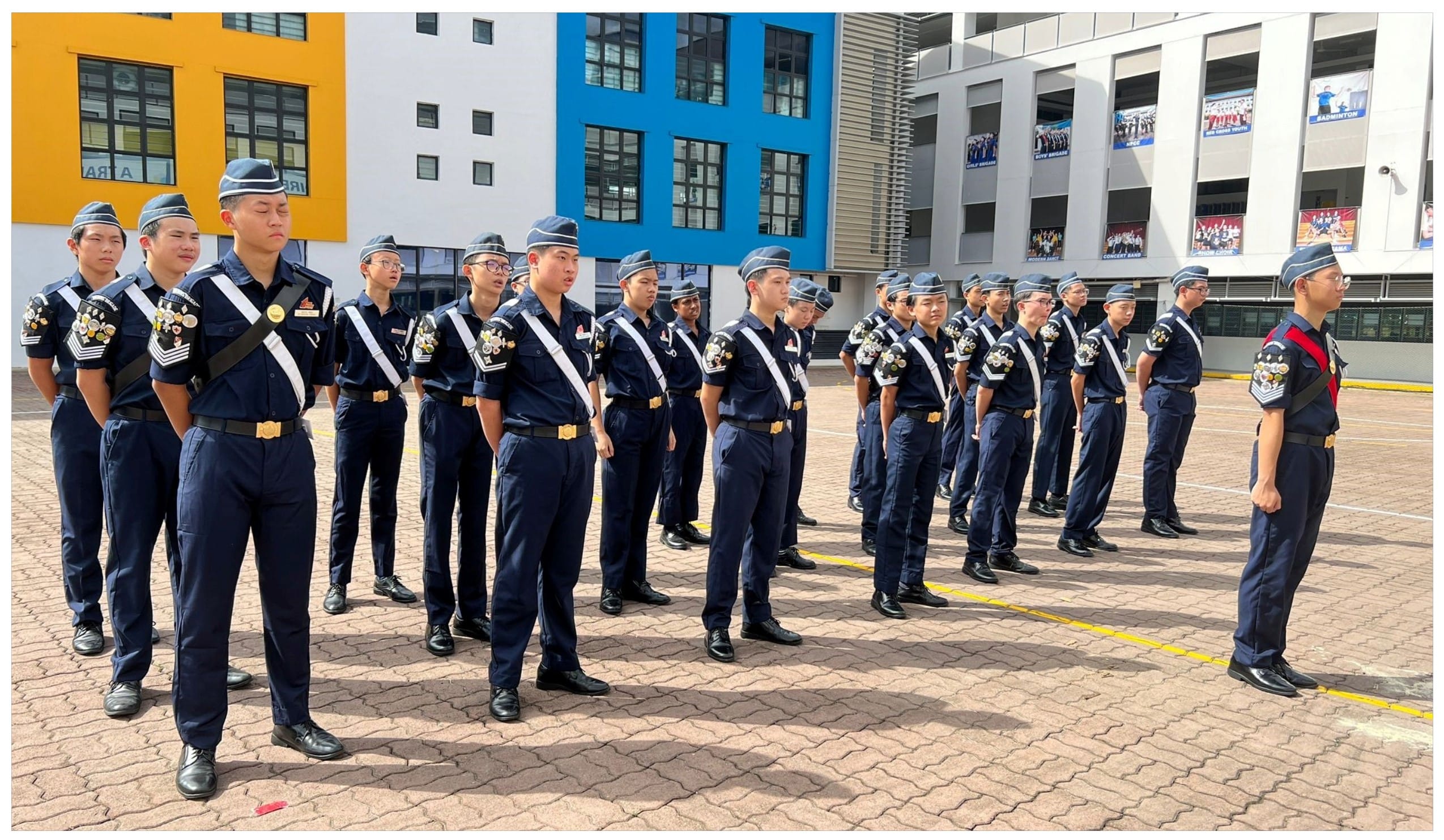 A group of young cadets in navy blue uniforms and caps stand in formation on a school courtyard.