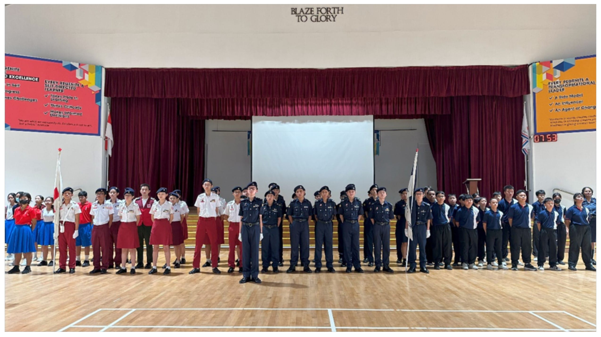 Students in various uniforms stand in rows in a school hall with a stage and "Blaze Forth To Glory" sign above.
