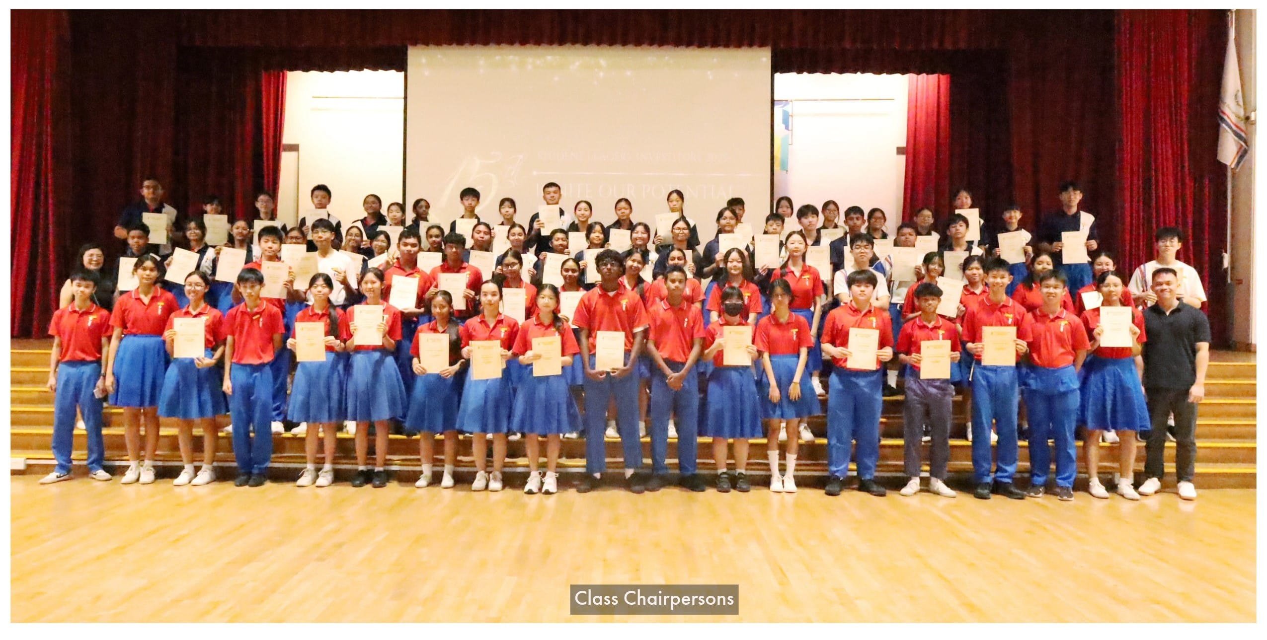Large group of students in red and blue uniforms holding certificates on a school stage, labeled Class Chairpersons.