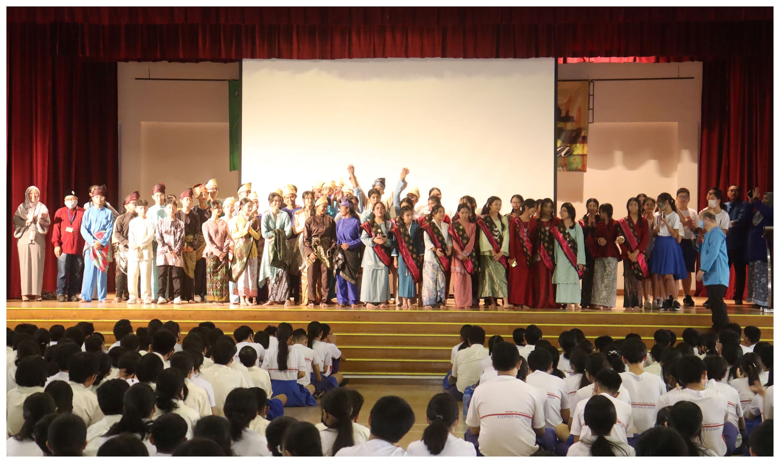 A large group in colorful traditional costumes stands on a school stage before a seated student audience in white uniforms.