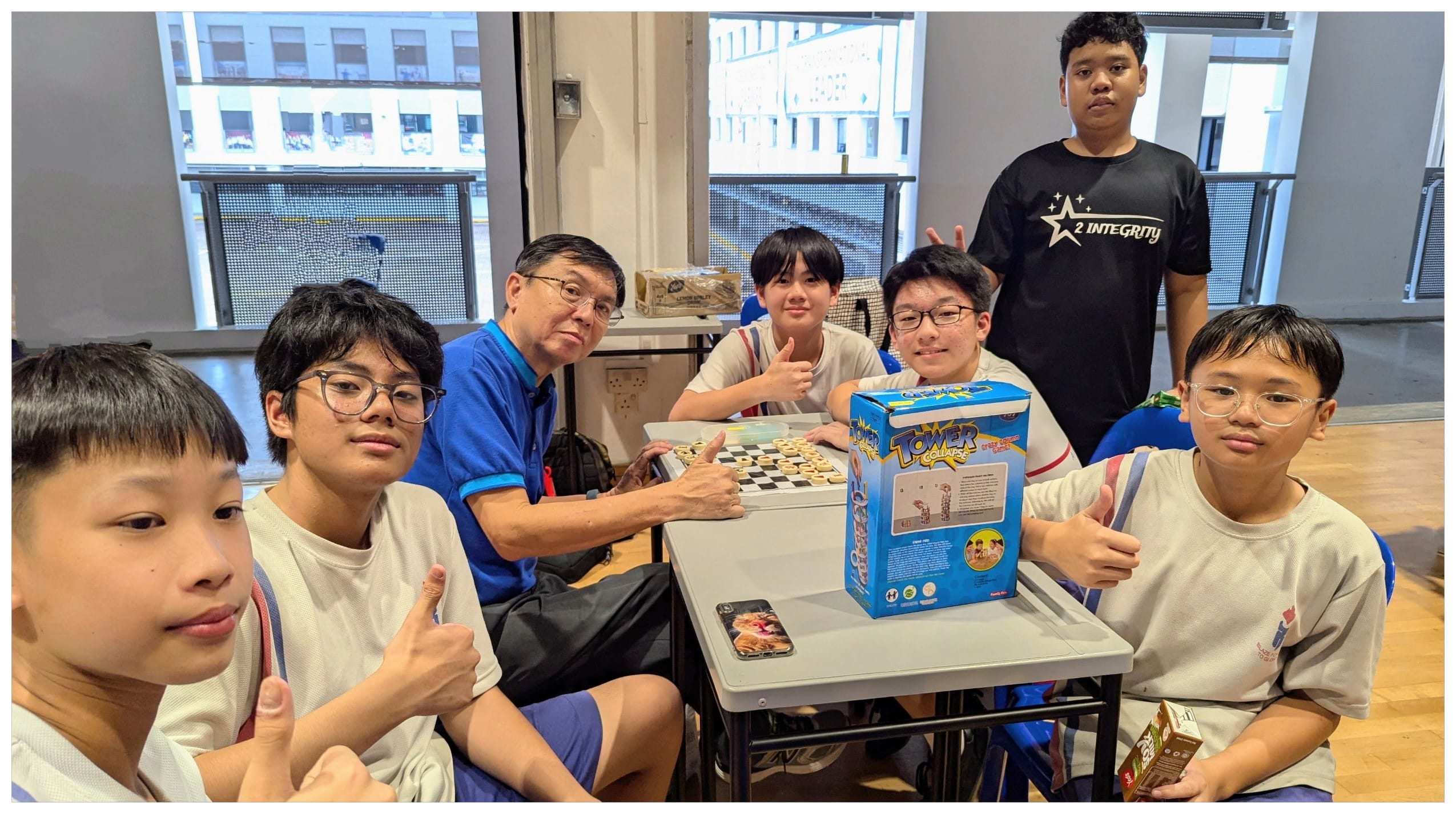Six youths and an older man pose around a table with a checkerboard and Tower Collapse board game box.