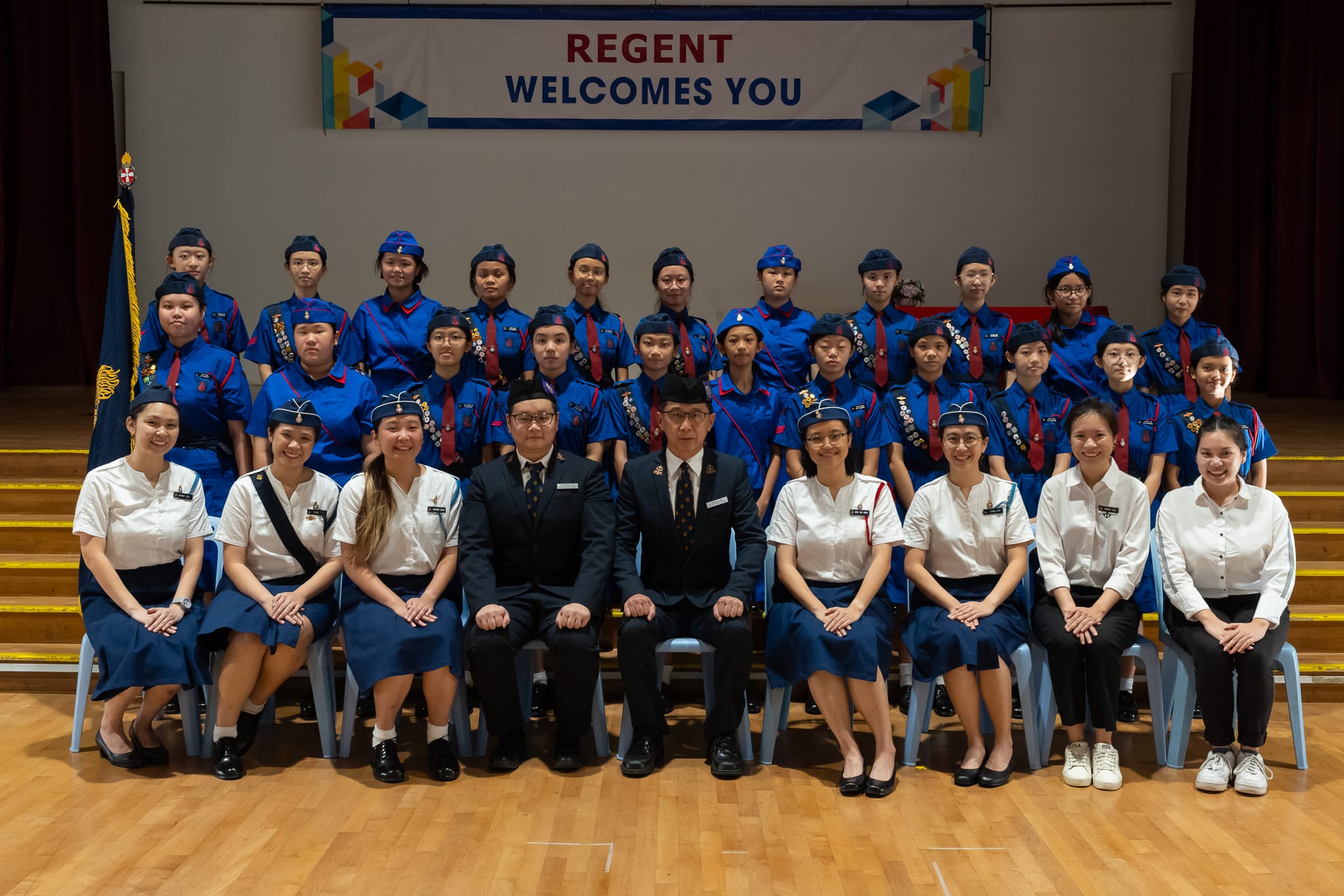 Group photo of uniformed students and staff in a hall with a banner reading Regent Welcomes You.