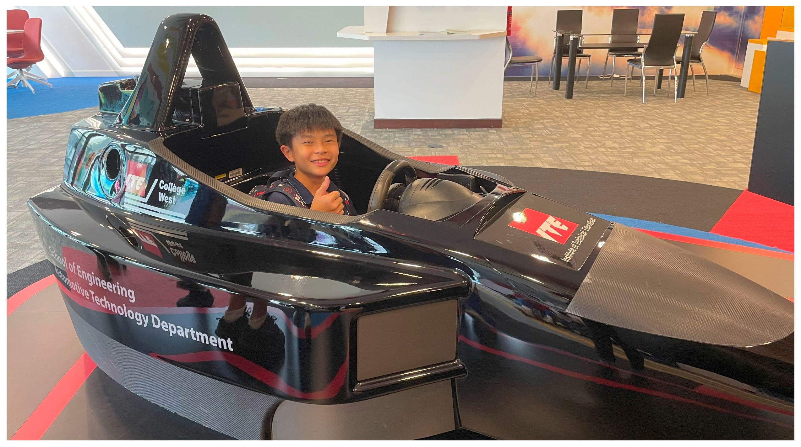 A smiling boy gives a thumbs up while sitting inside a black Formula 1 style racing car simulator indoors.