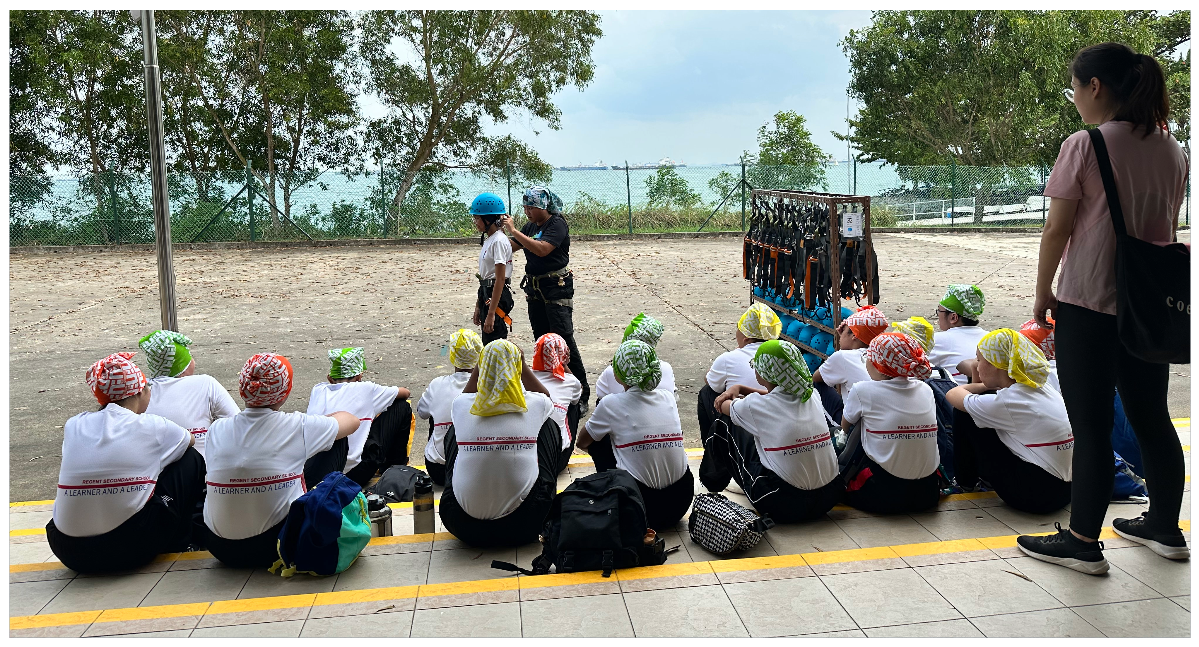 Students in white shirts sit on the ground watching an instructor fit a helmet on a student outdoors near water.