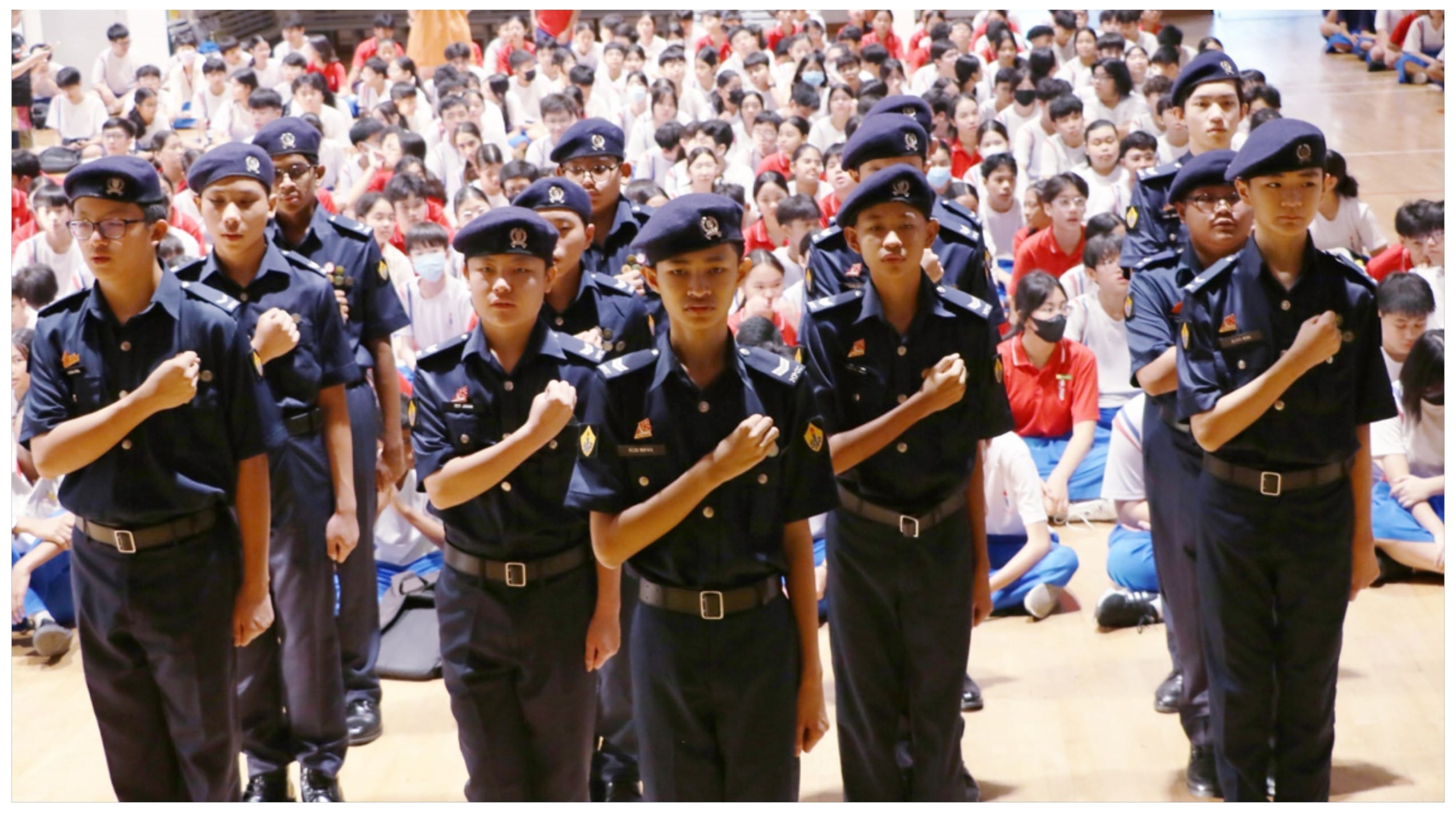 Young cadets in navy uniforms and berets march with fists raised before a large seated student audience.