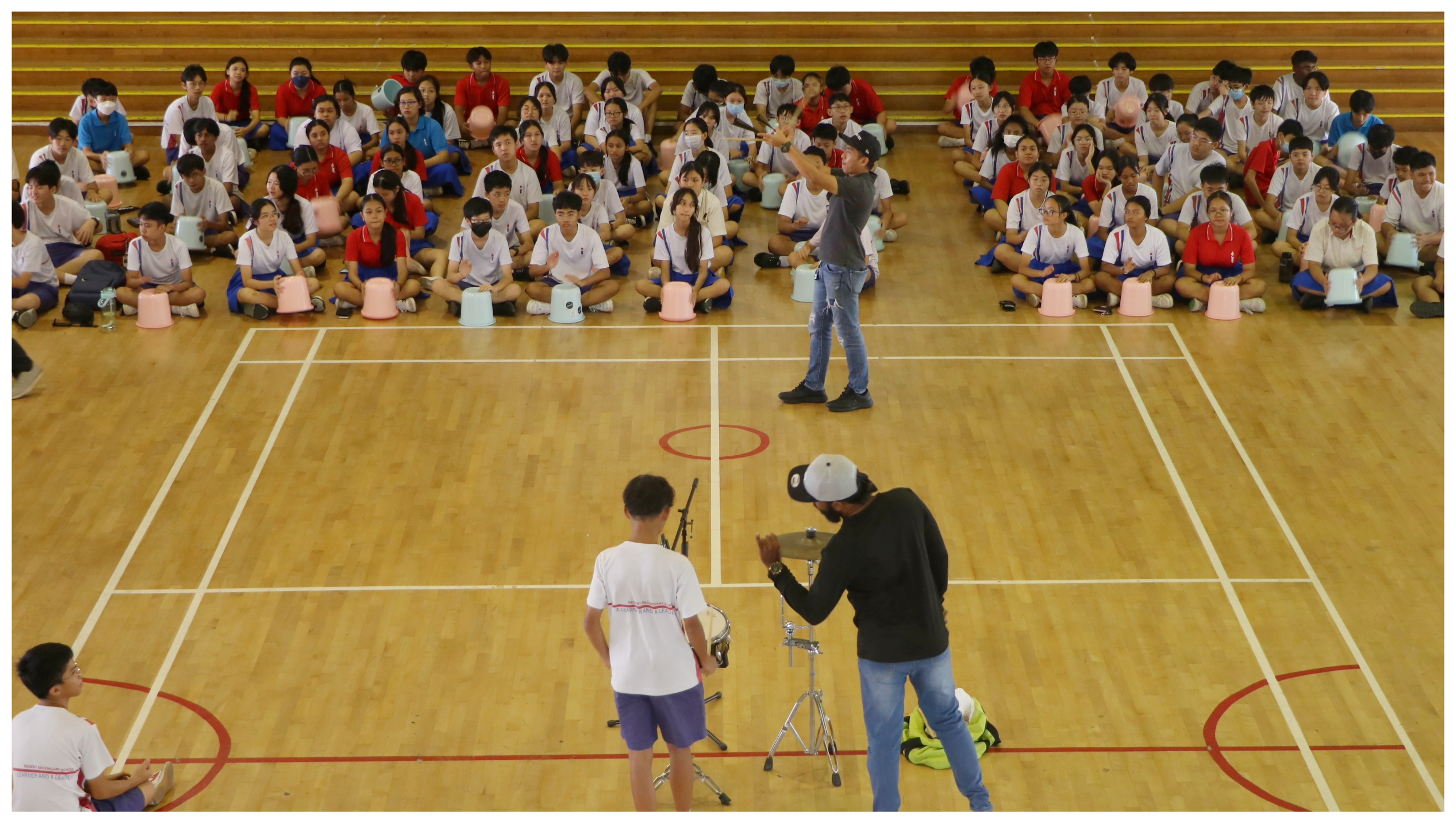 Students sit on a gym floor with buckets as drums while two adults and a teen lead a rhythm lesson.