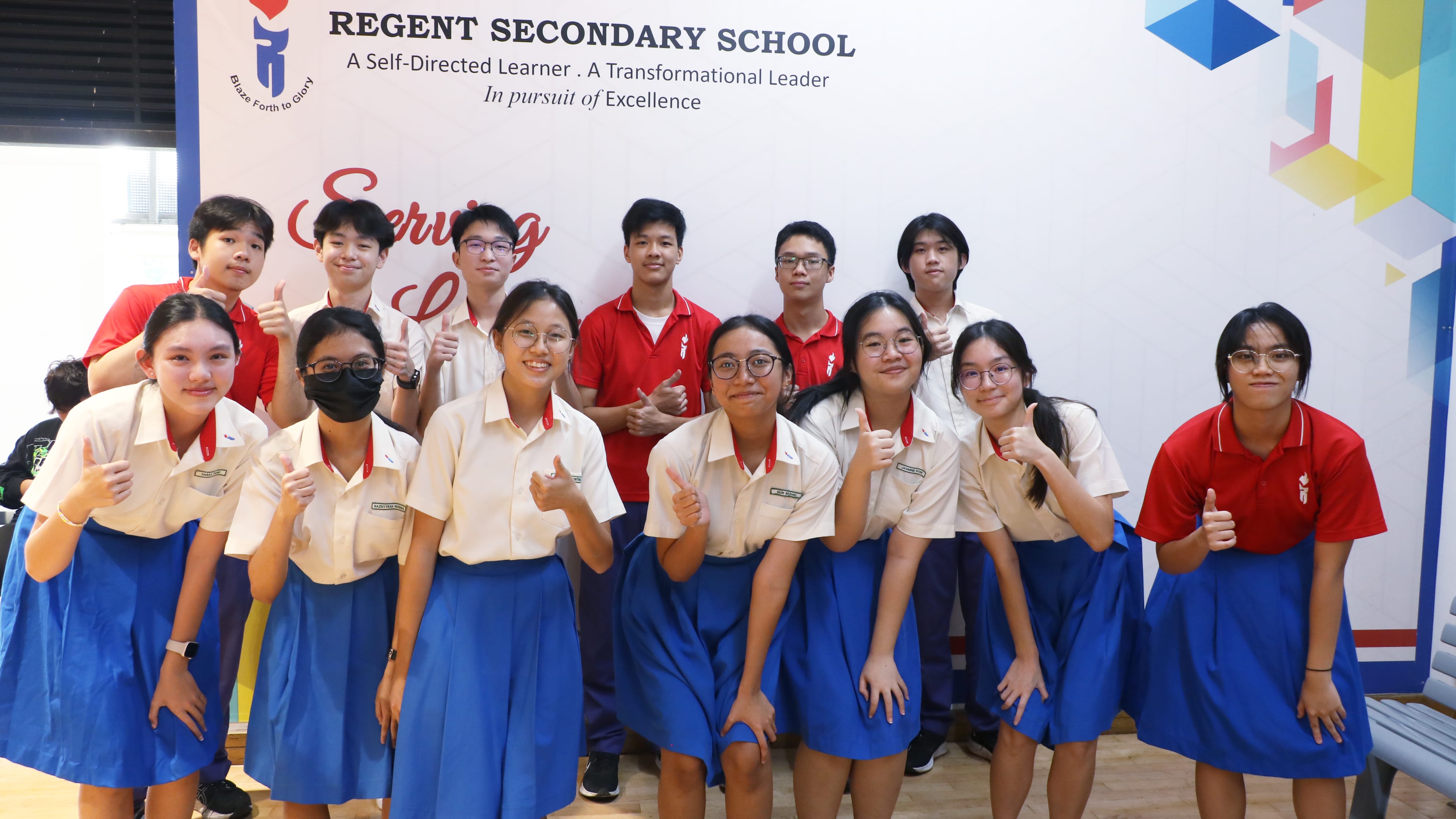 Group of students in school uniforms posing with thumbs up in front of a Regent Secondary School banner.