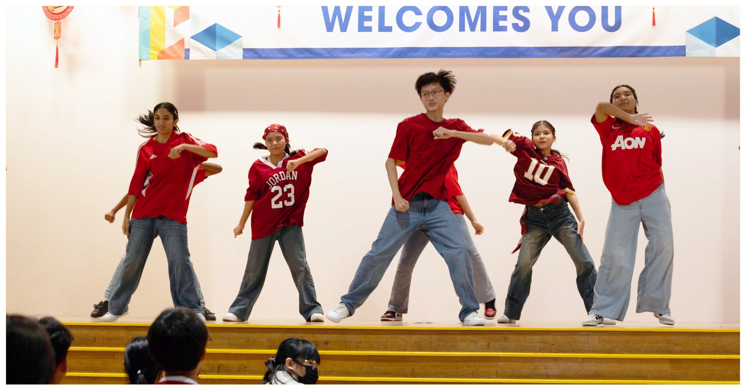 Five kids in red jerseys and jeans perform a dance on a school stage under a "Welcomes You" banner.