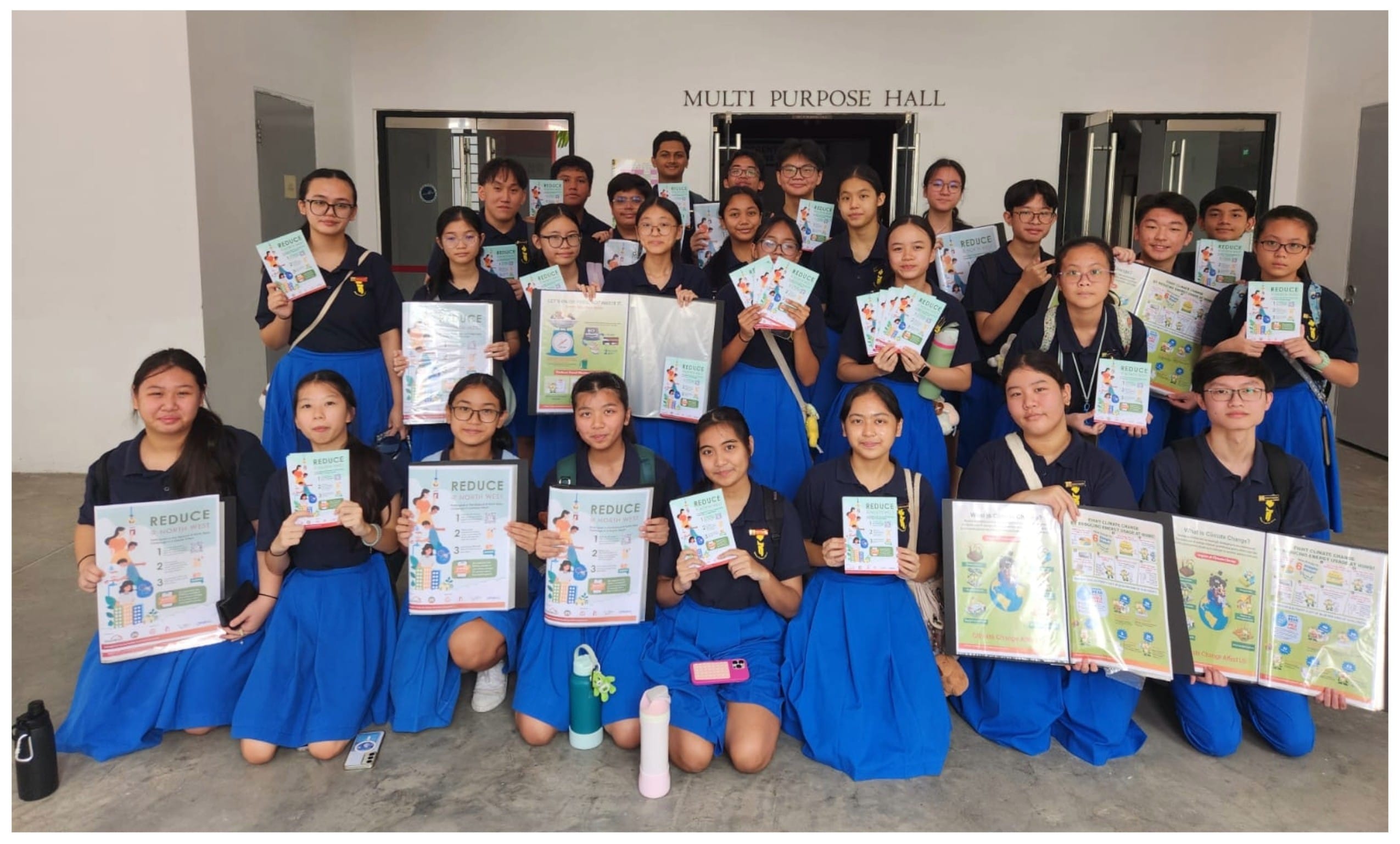 Students in blue uniforms hold up Reduce-themed posters in front of a Multi Purpose Hall entrance.