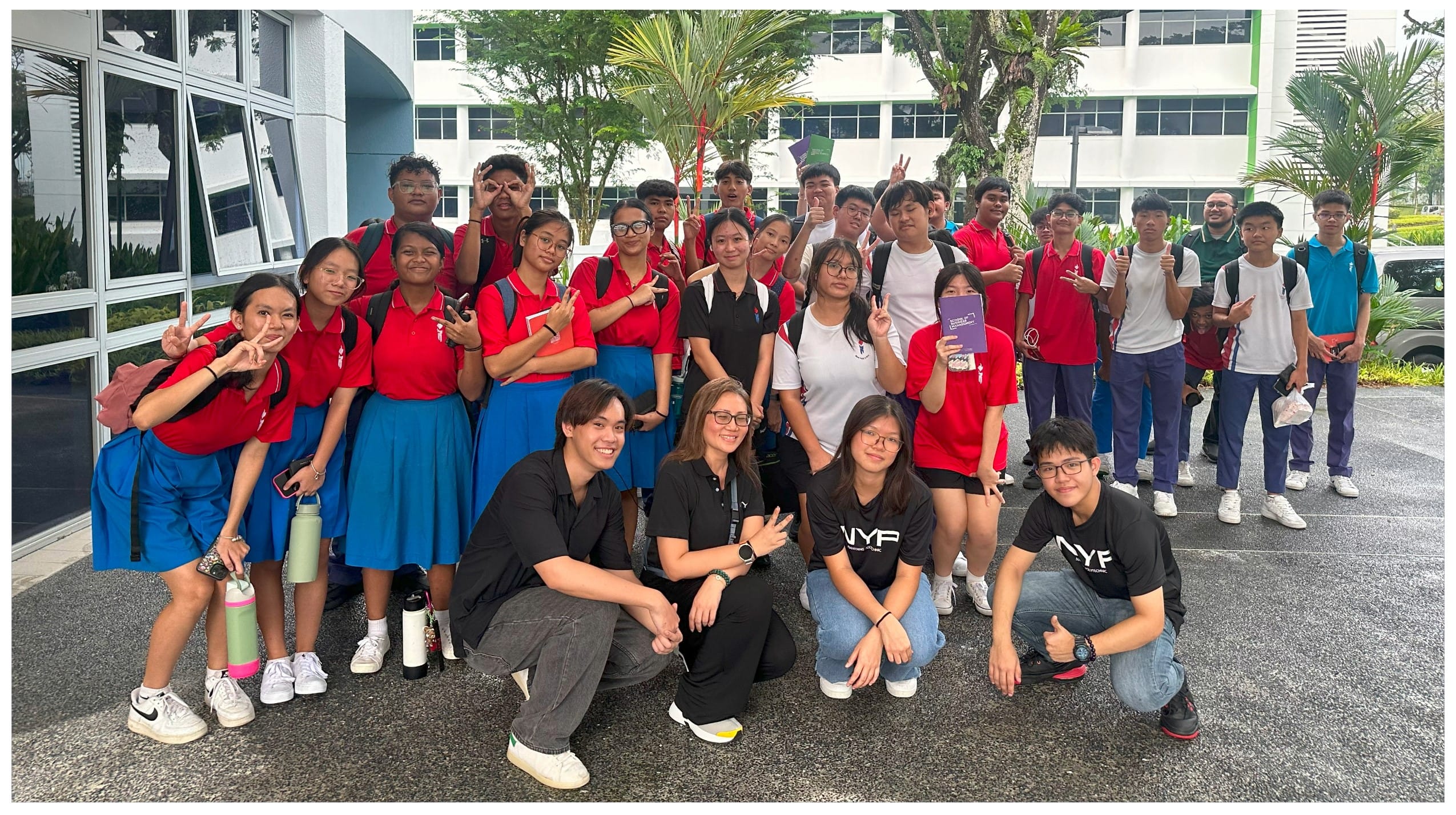 A large group of students in red uniforms and adults in black shirts pose outdoors near a white building.