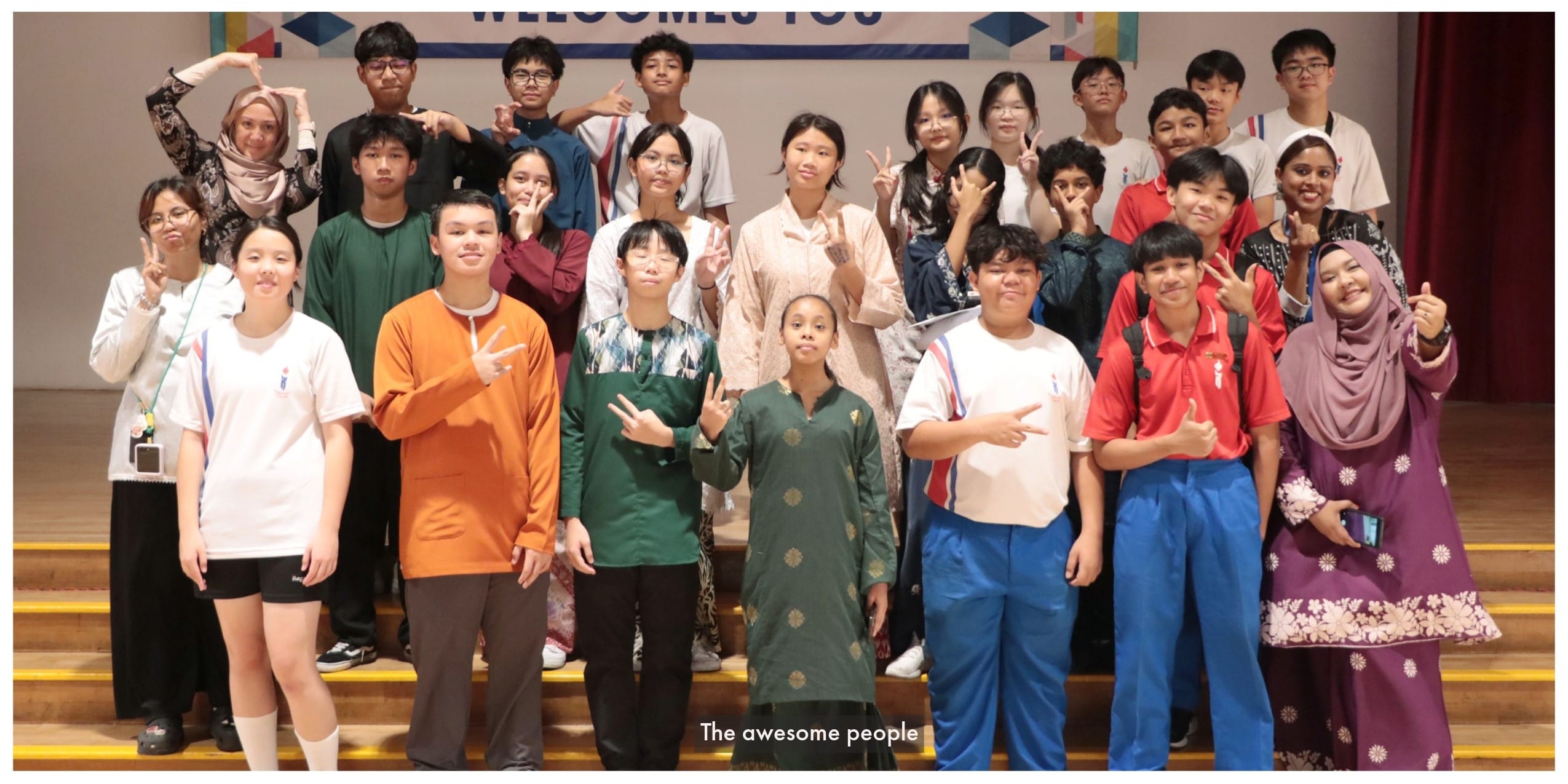 A large group of students and adults posing with peace signs in front of a welcome banner on a stage.