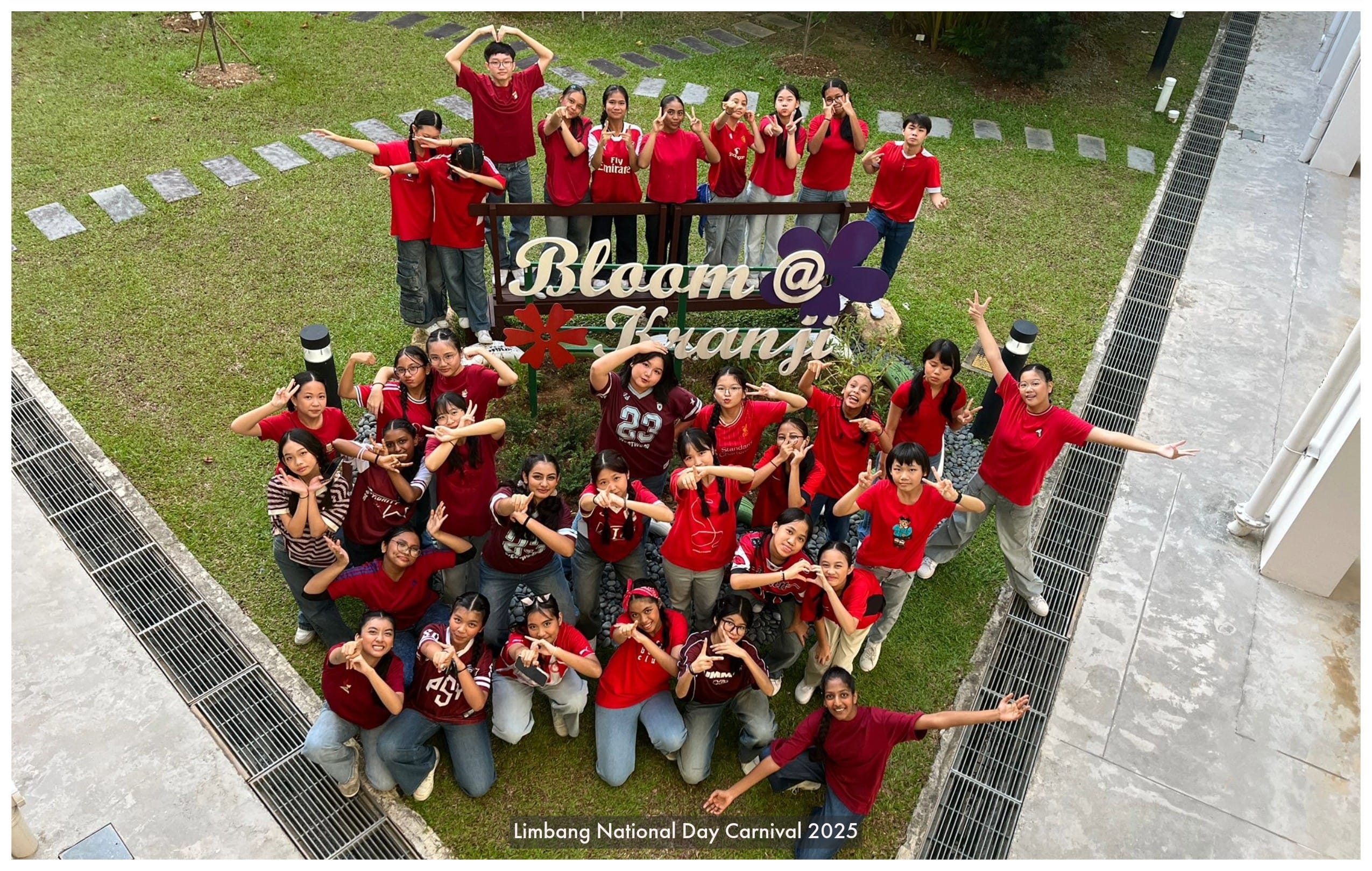 A large group of students in red shirts pose around a Bloom @ Kranji sign at Limbang National Day Carnival 2025.
