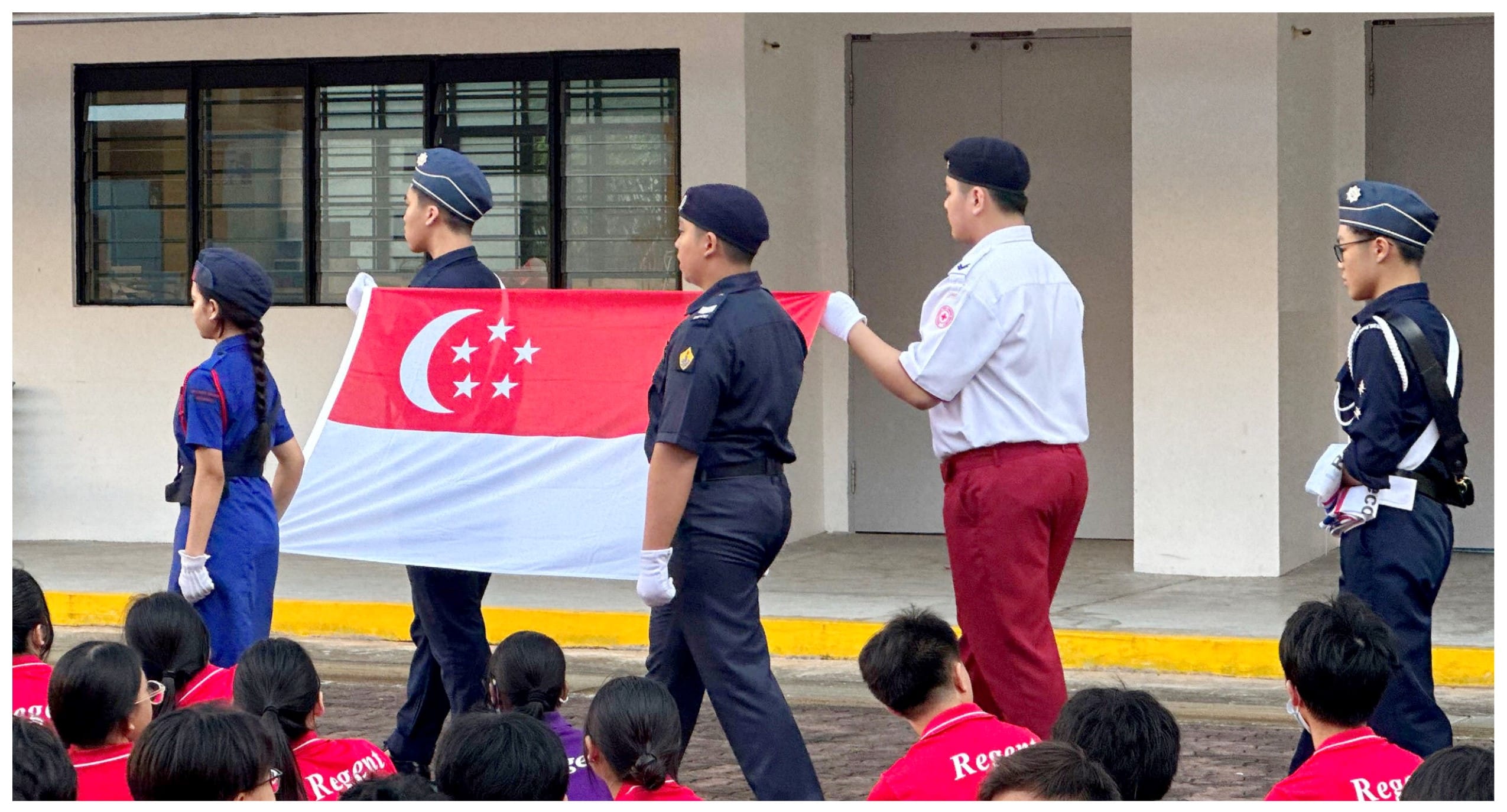 Students in uniform carry the Singapore flag during a school parade ceremony before a seated crowd.