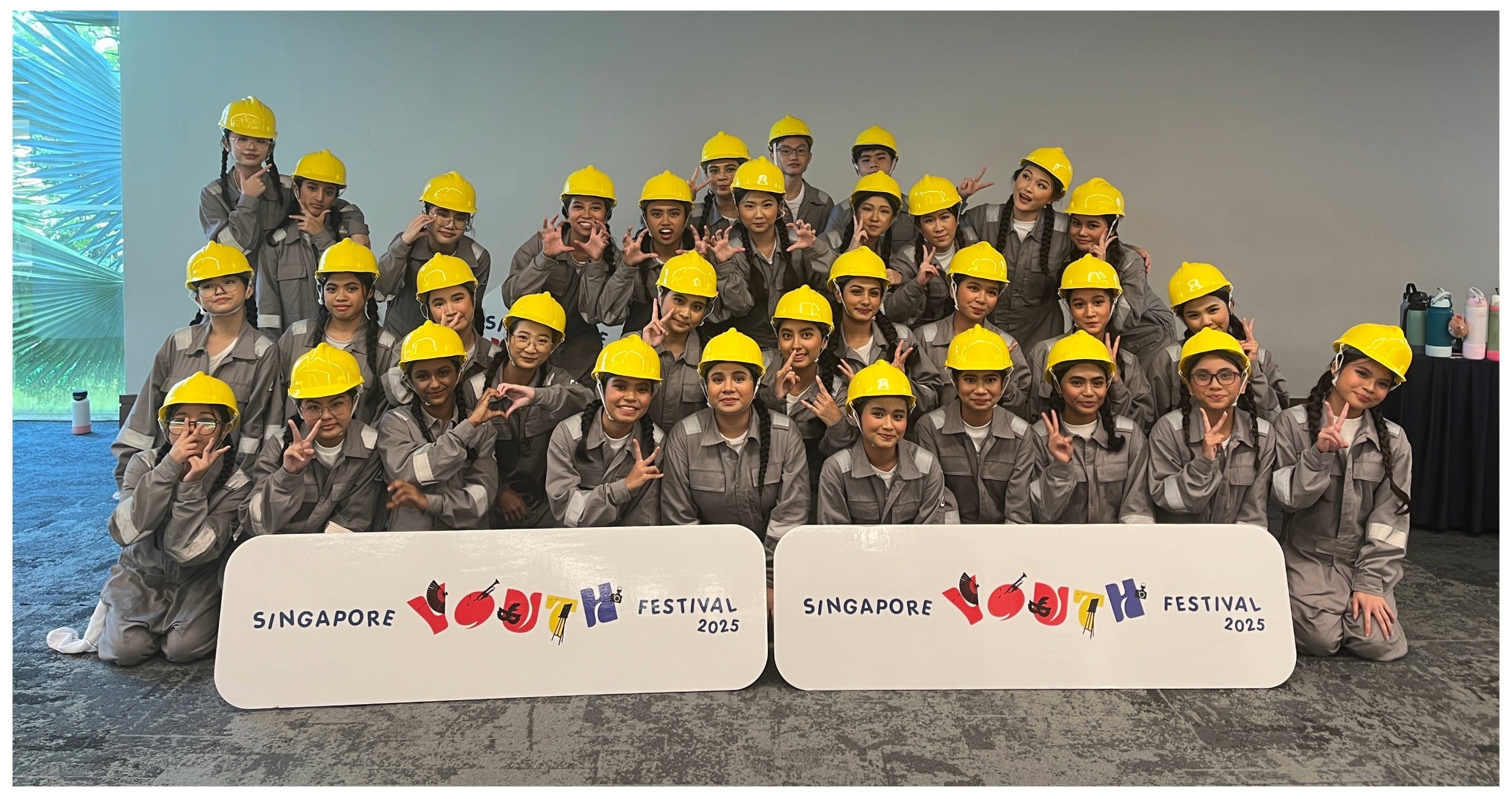 A large group in gray coveralls and yellow hard hats poses with Singapore Youth Festival 2025 signs.