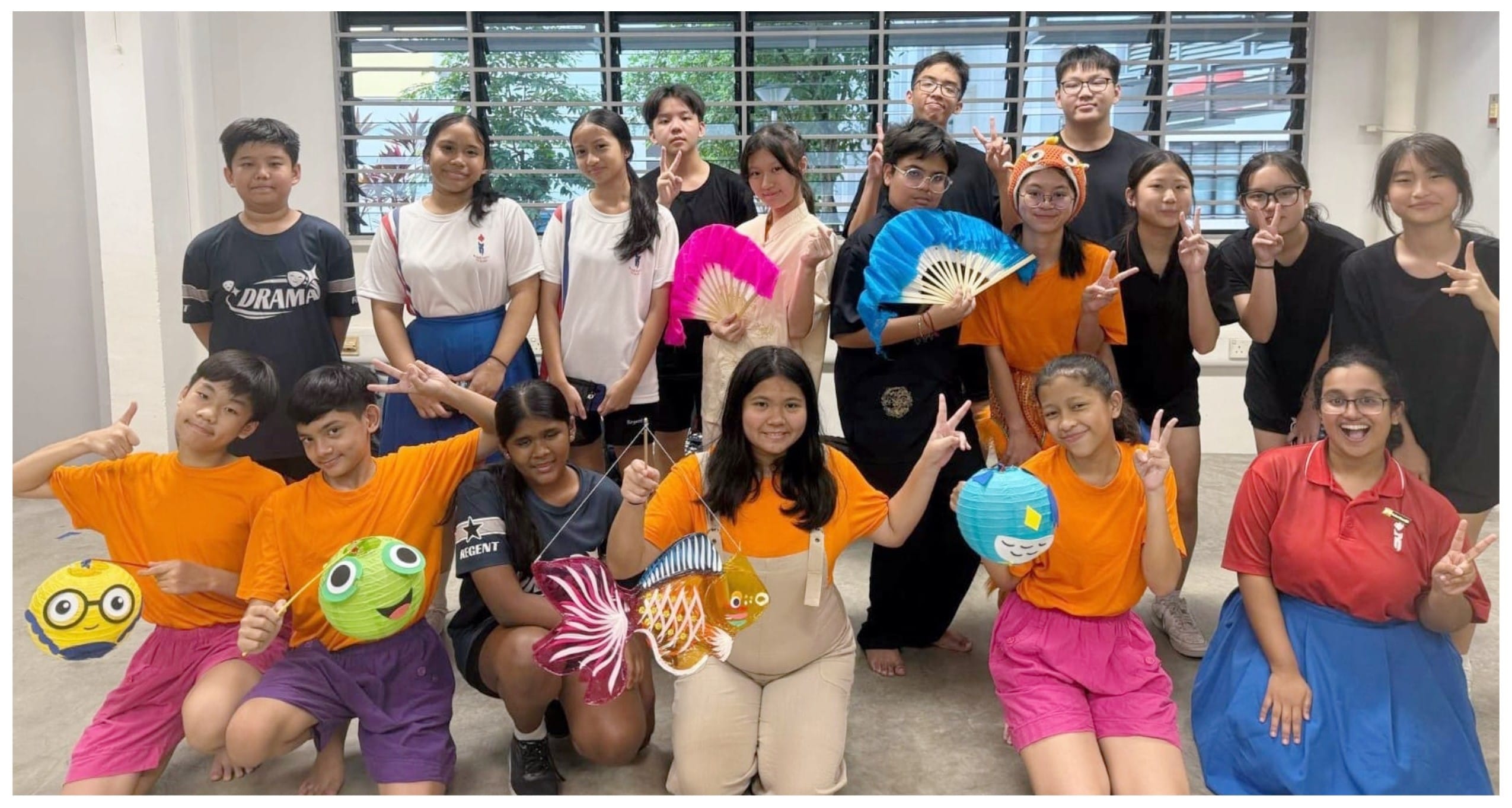 Group of students posing indoors, holding colorful paper lanterns and decorative fans, some making peace signs.
